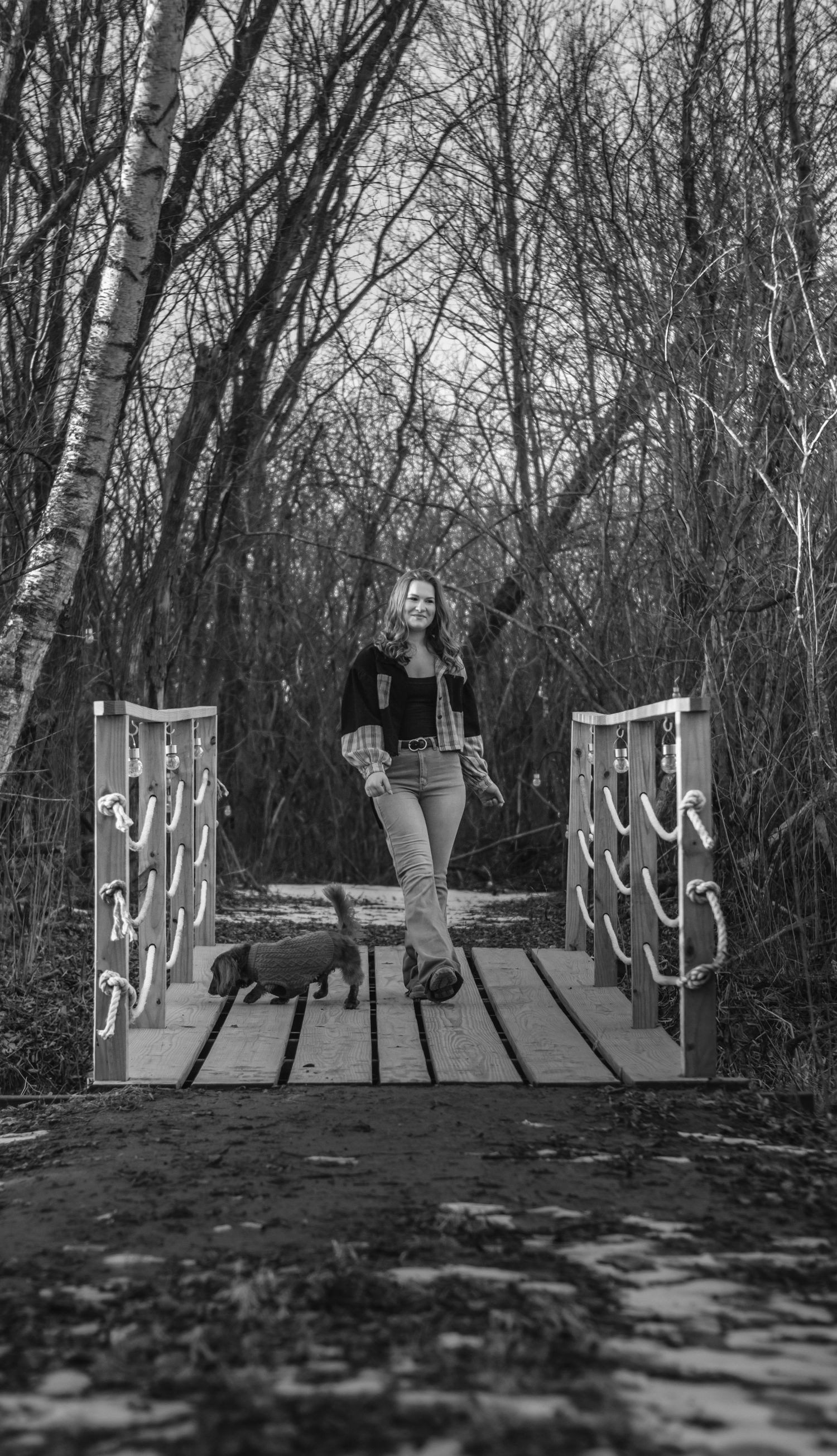 Woman smiling on wooden bridge with dog in black and white forest setting.