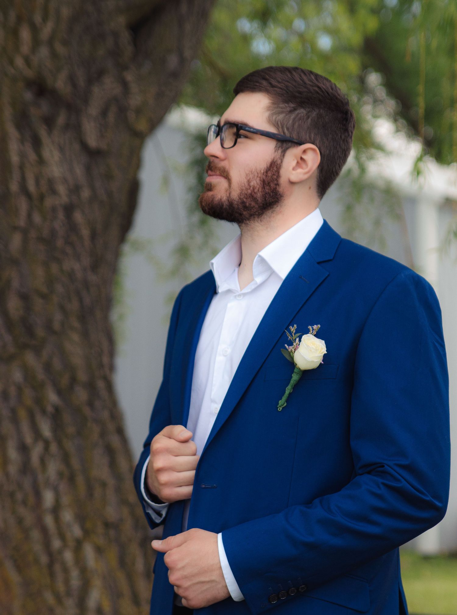 A person wearing a navy blue suit and white shirt adjusts their jacket collar while standing outdoors near trees.