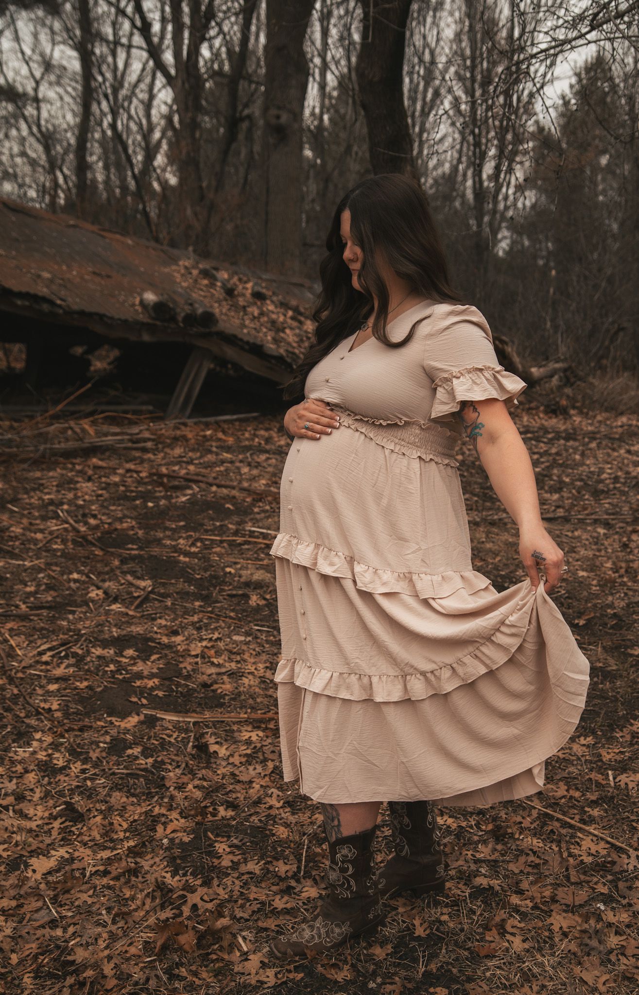 Pregnant woman in beige ruffled dress and boots holding her belly in an autumn woodland setting.