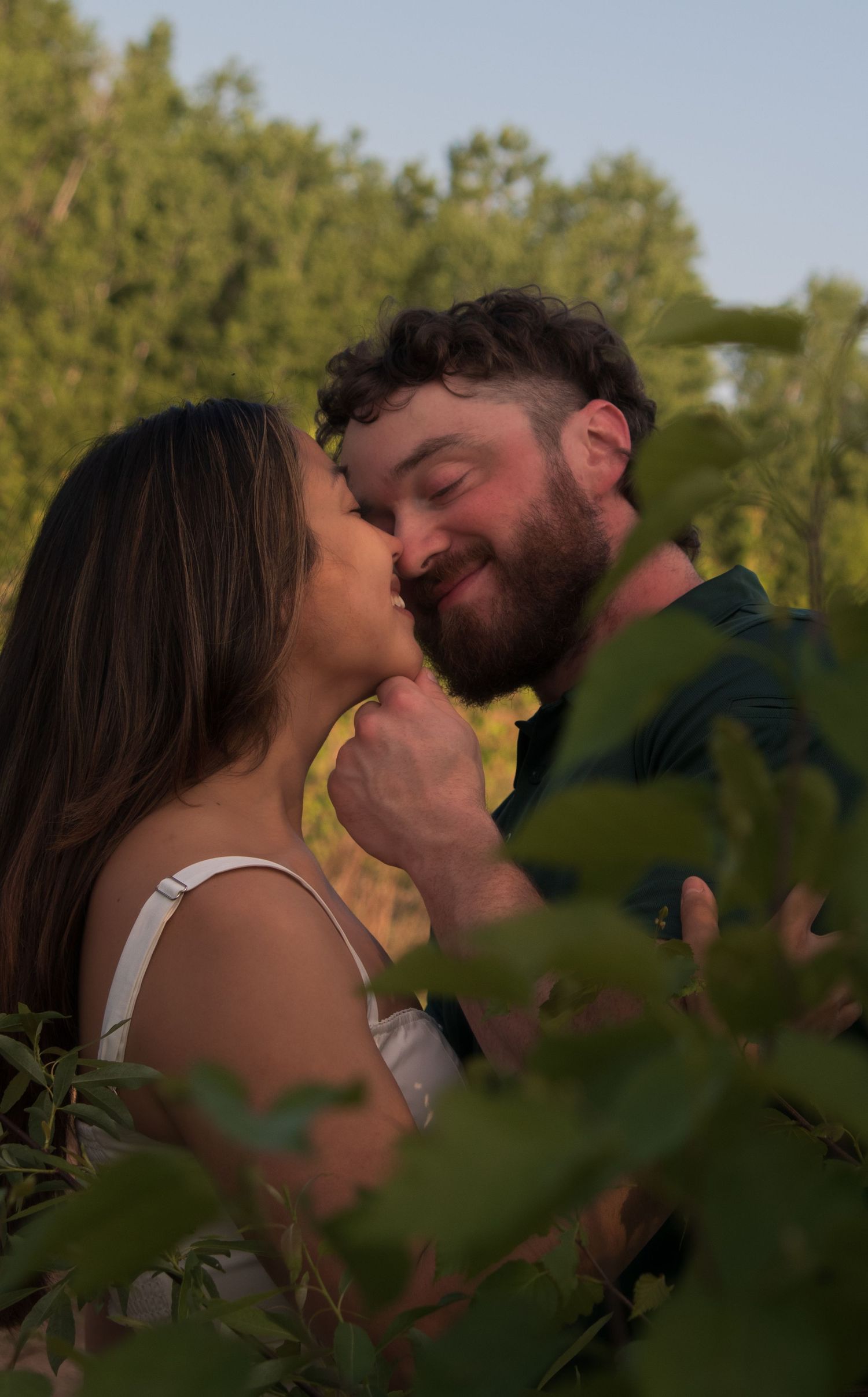 A romantic couple shares an intimate moment among green foliage at sunset.