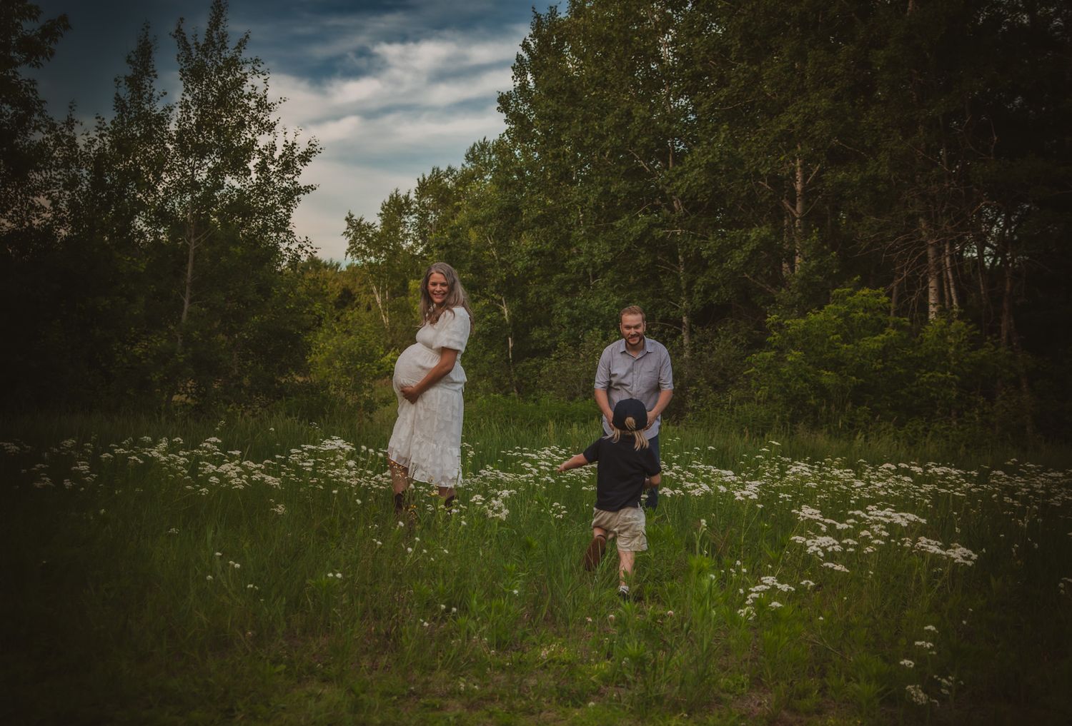 Pregnant woman in white dress with husband and child playing in wildflower meadow surrounded by lush green trees.
