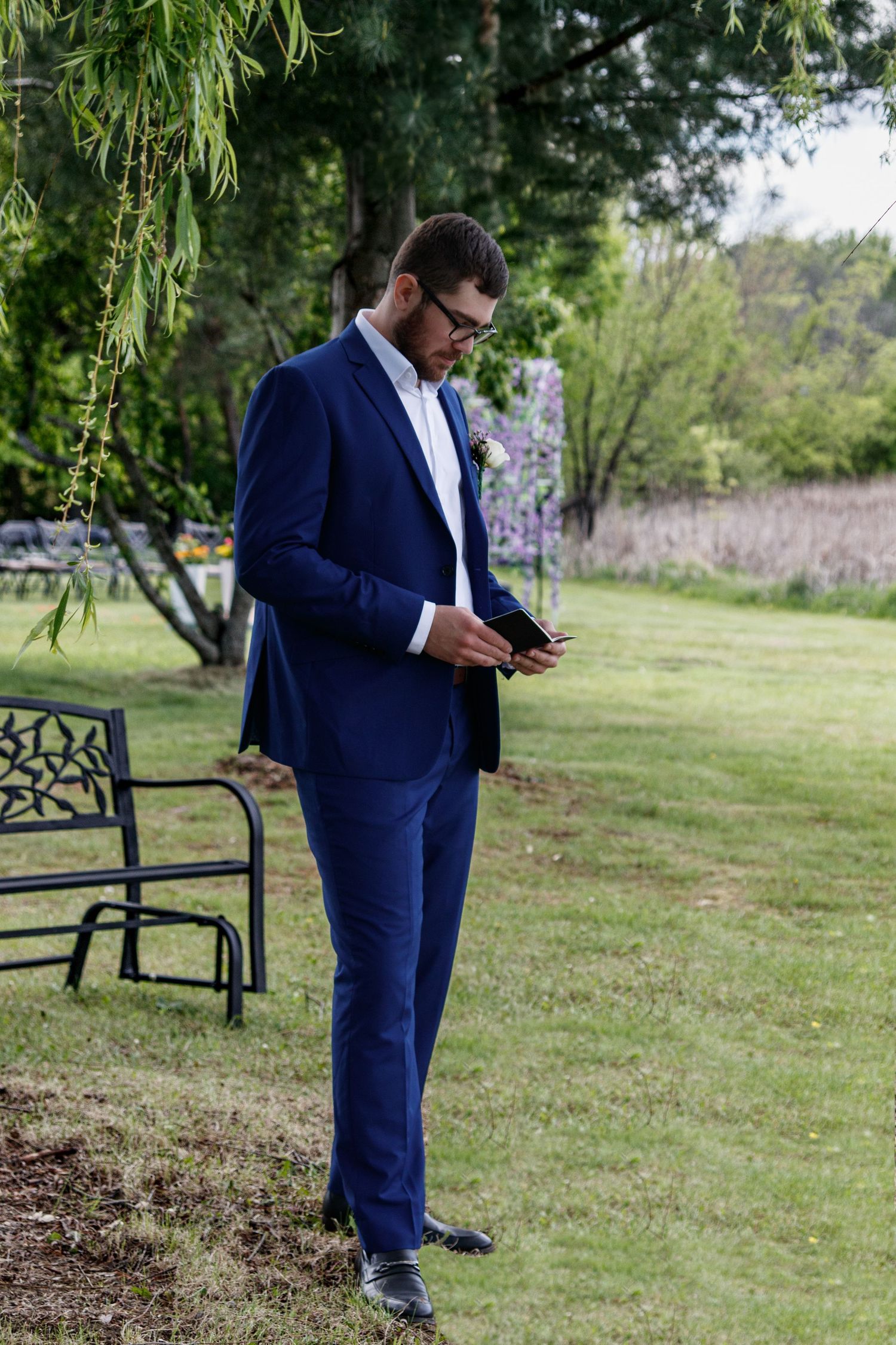 A person in a navy blue suit stands near a decorative black metal bench in a lush outdoor garden setting.