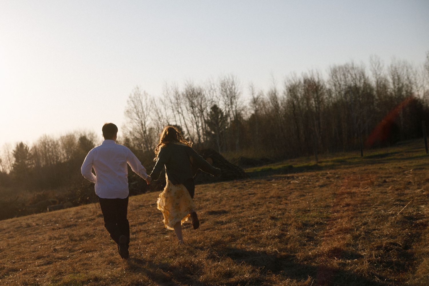 Couple holding hands running through a sunlit field at golden hour with trees in the background.