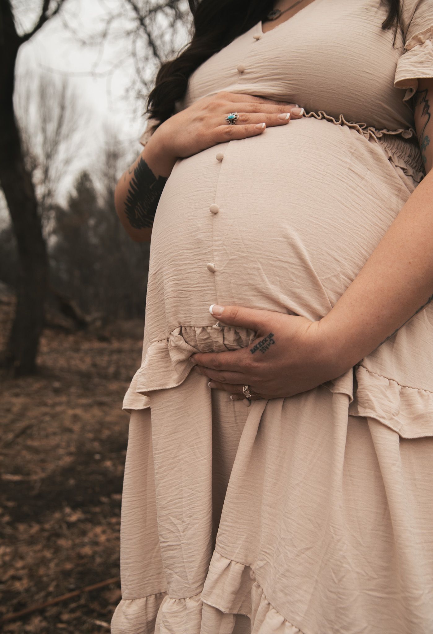 Pregnant woman in blush ruffle dress holding baby bump with tattooed hands in autumn setting.