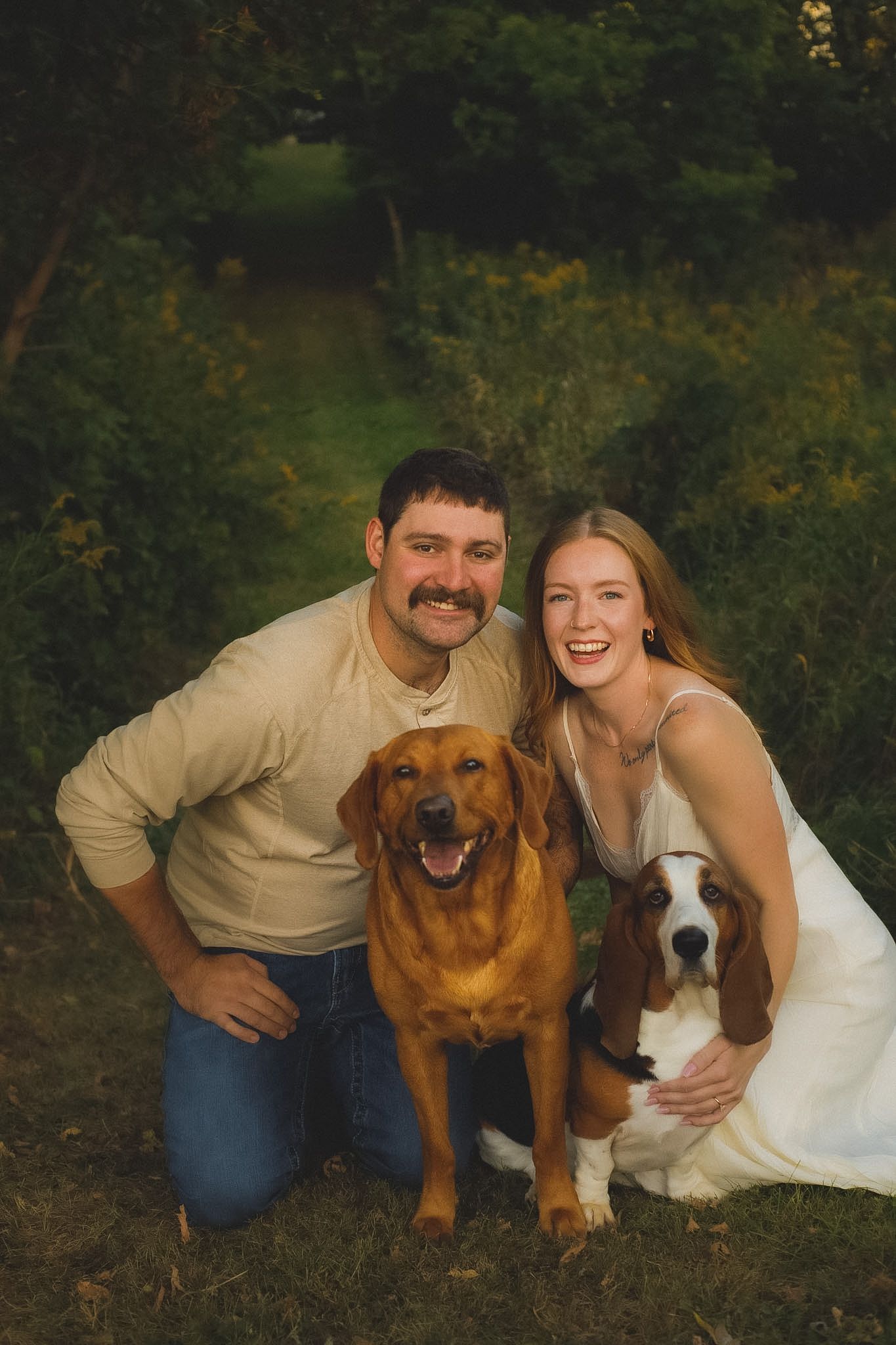 A smiling couple kneels outdoors with a golden retriever and basset hound, surrounded by lush green trees.
