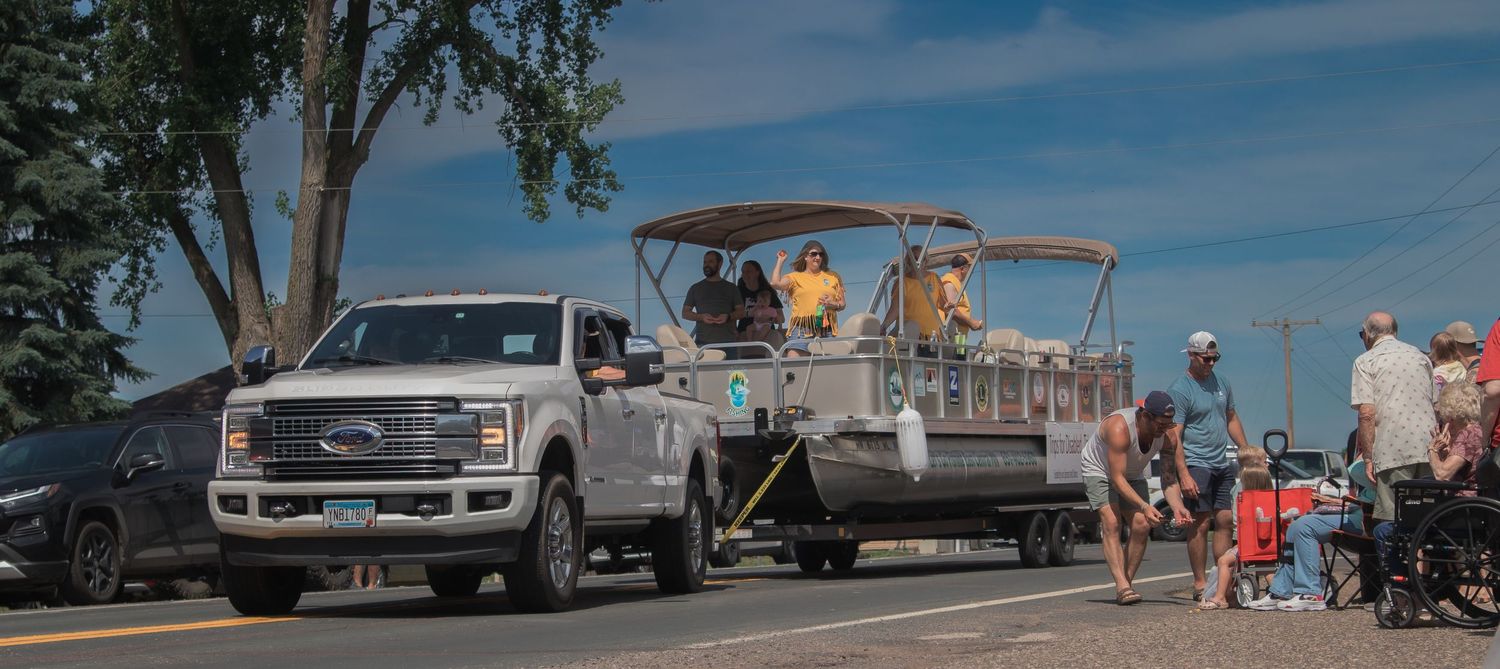 A panoramic view of a lakeside gathering with boats, trucks, and people on a sunny summer day.