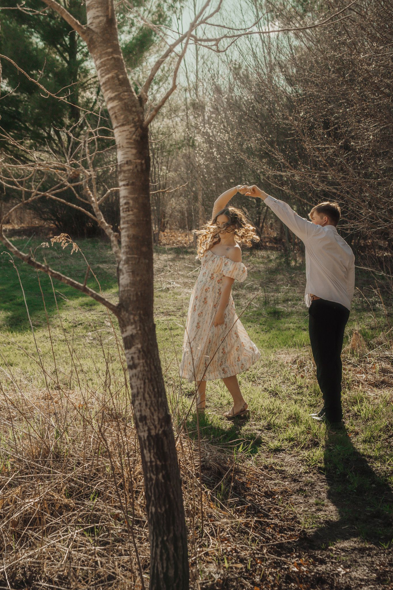 Couple dancing joyfully in a sunlit forest, woman in floral dress twirling as man in white shirt leads.