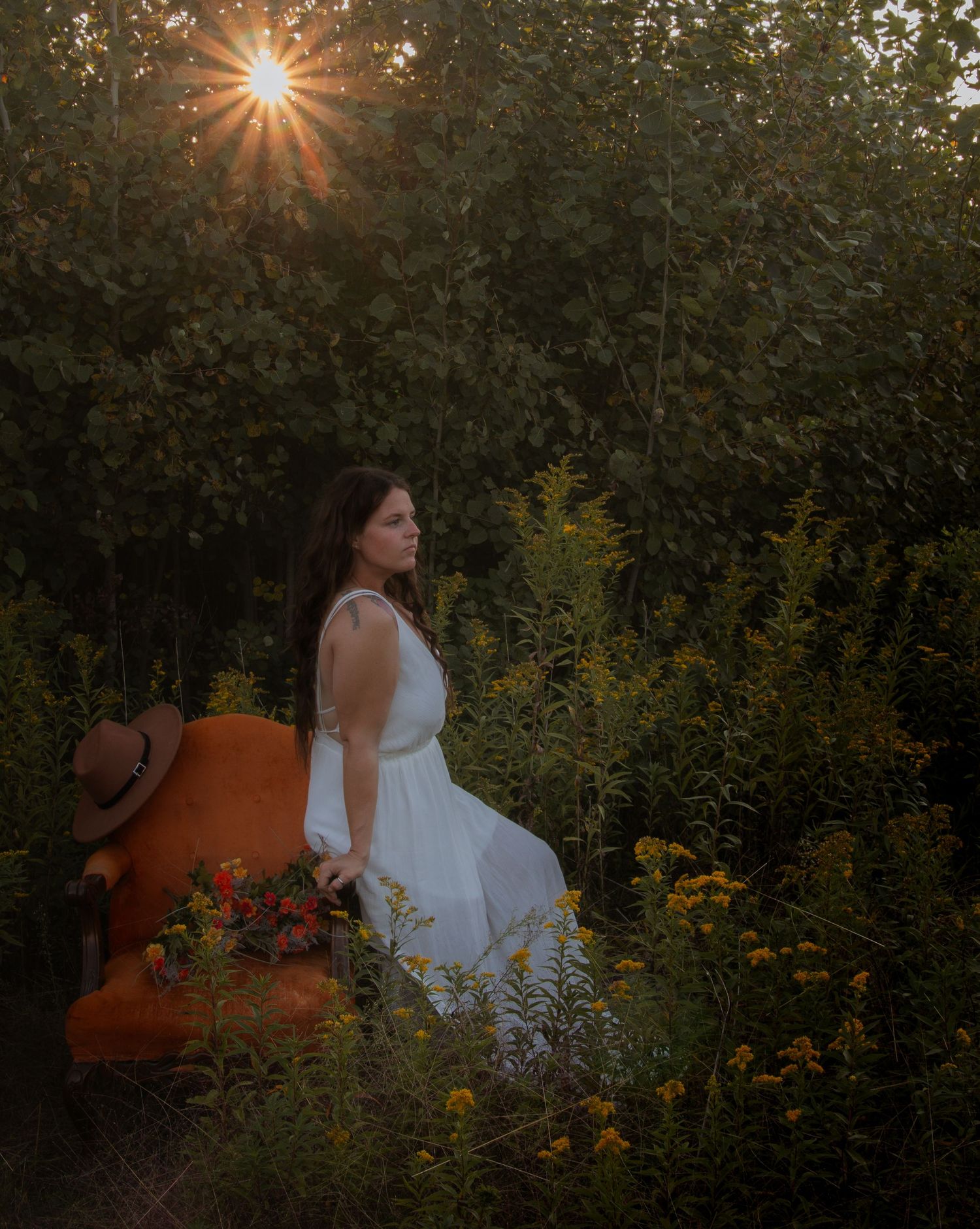 A white dress adorned figure sits on an orange vintage chair in a garden setting with sunburst through trees.