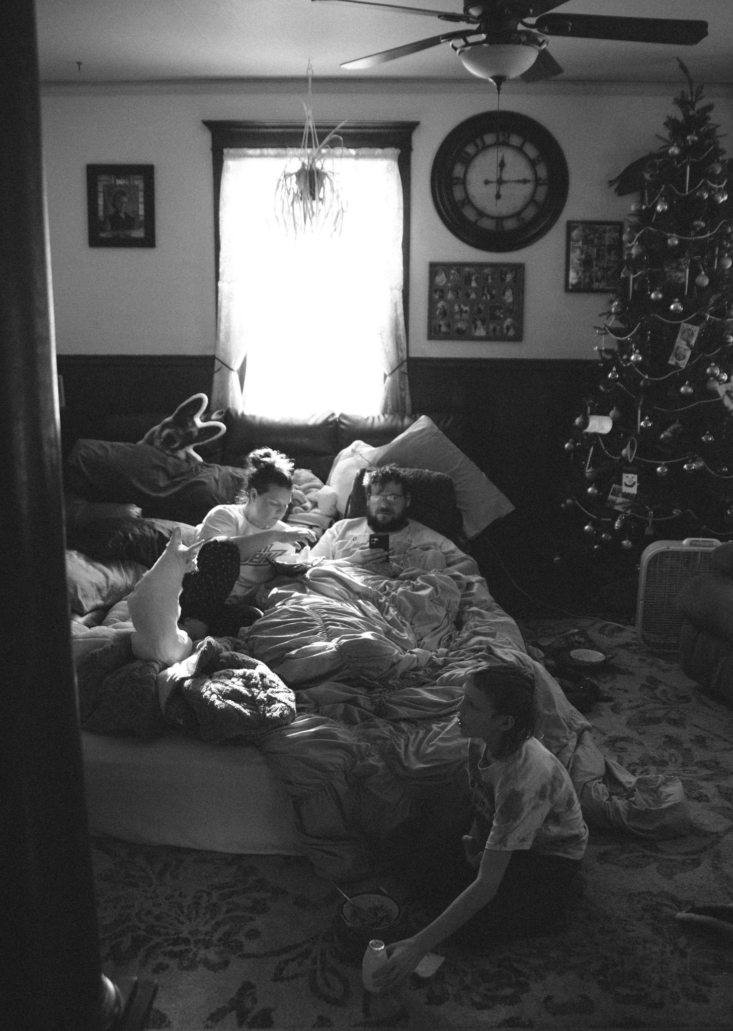 Black and white photo of family relaxing on bed near Christmas tree with clock on wall and ceiling fan.