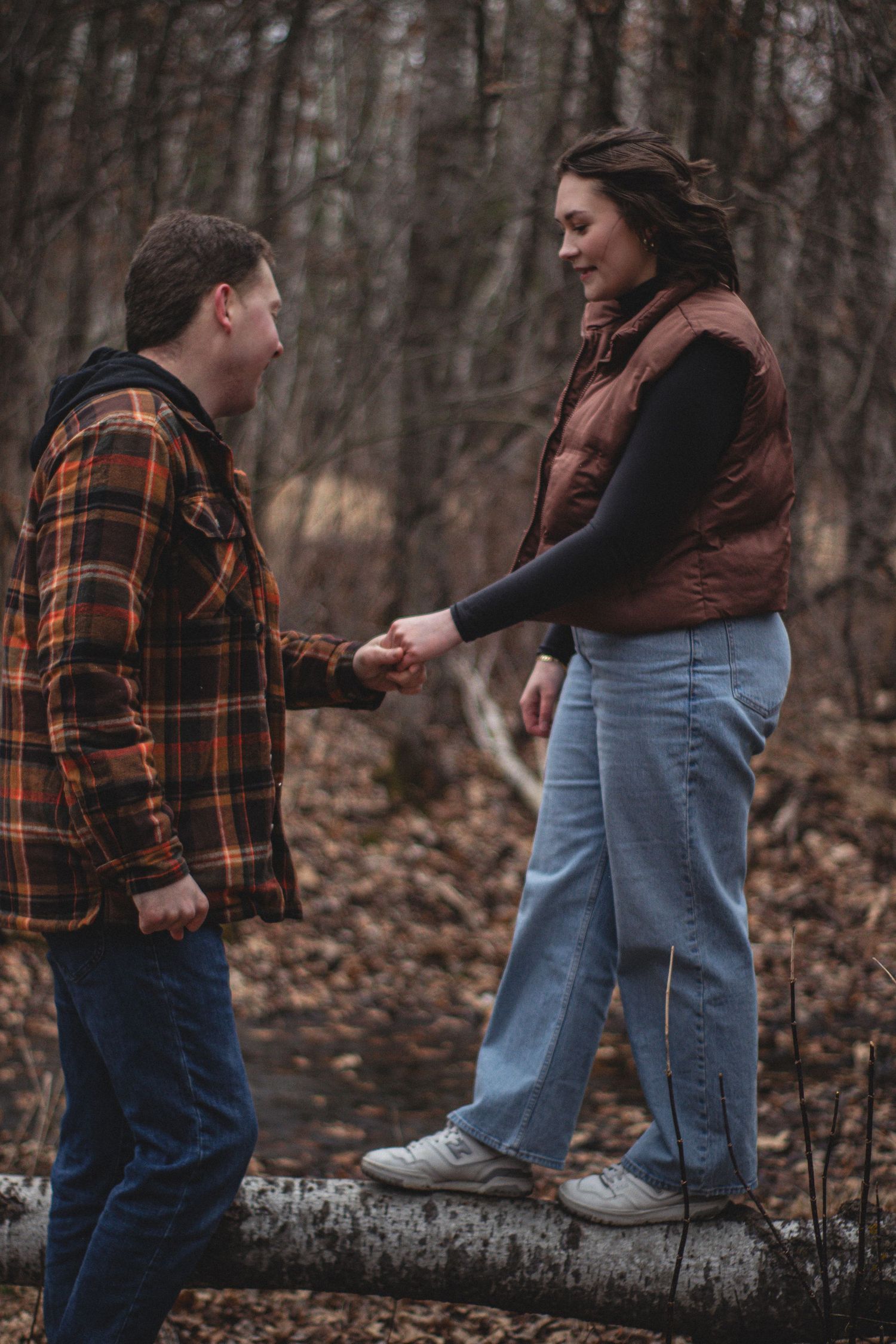 Two people in casual clothing play and dance together in a wooded area with fallen autumn leaves.