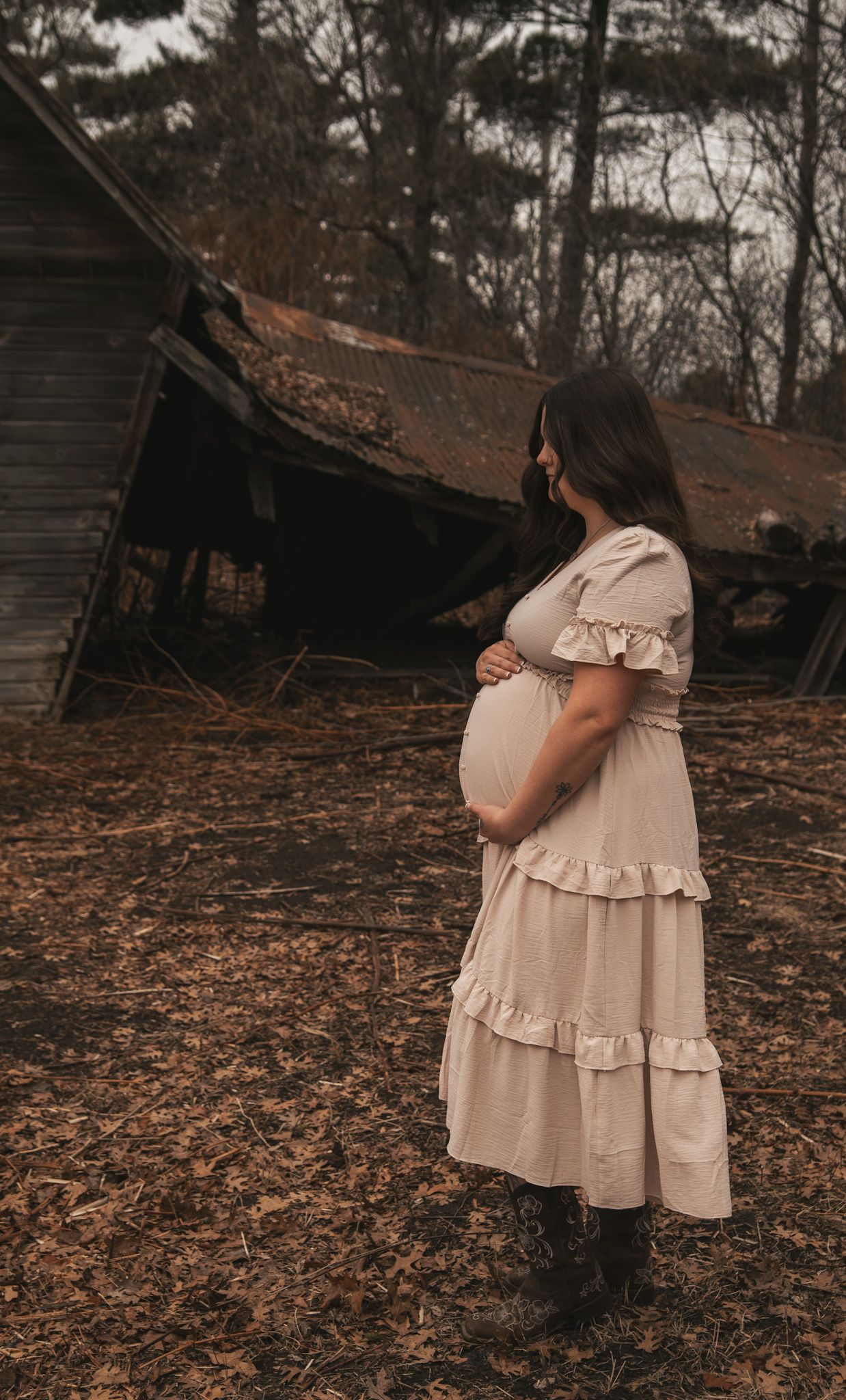 Pregnant woman in cream dress and boots cradling belly outdoors near rustic abandoned barn in autumn.