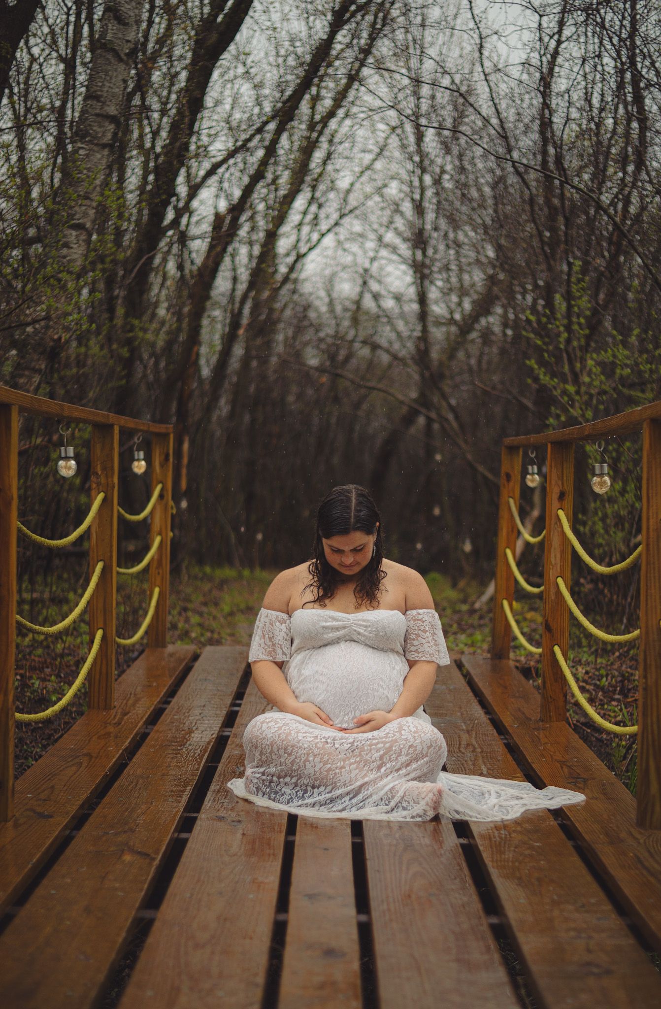 Pregnant woman in white lace dress sitting on wooden bridge surrounded by bare trees in a serene maternity photo.