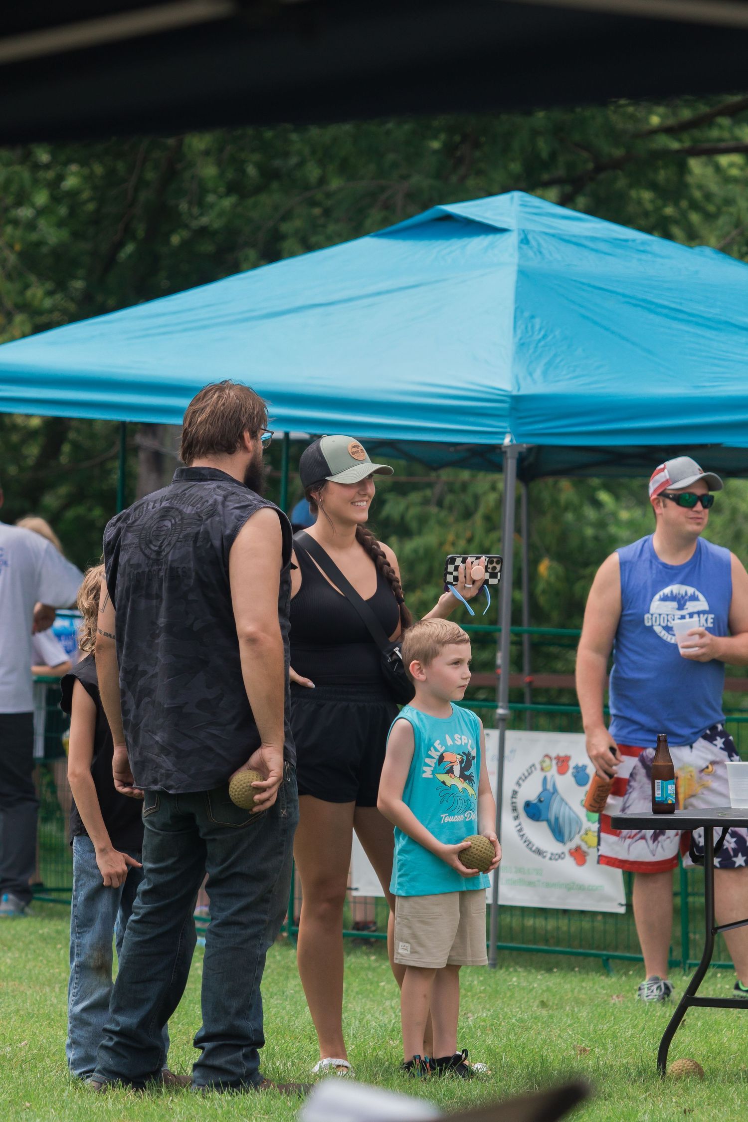 People gather under a blue pop-up tent at an outdoor event with tables and refreshments.