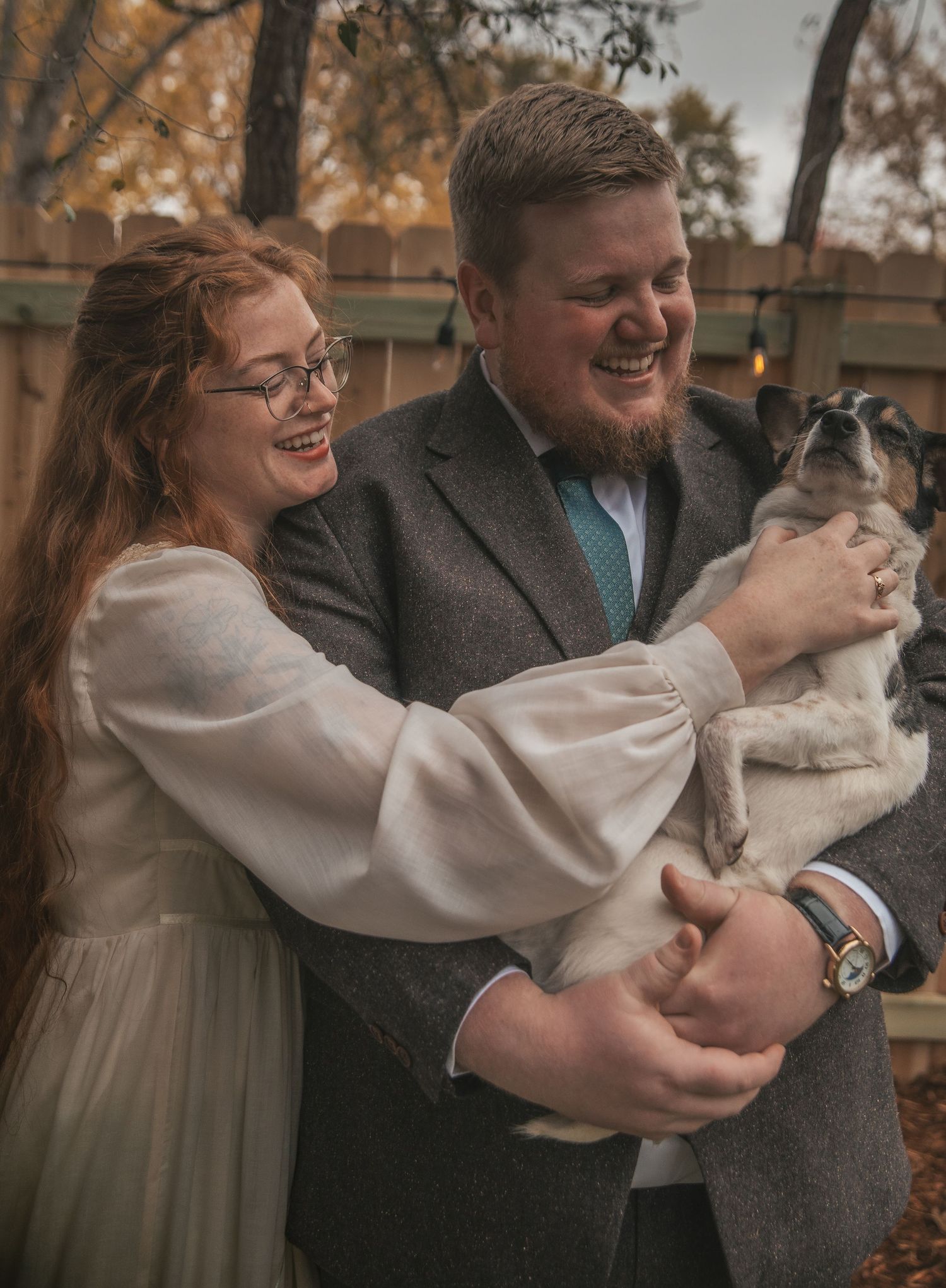 A couple shares a joyful moment outdoors while holding their small dog in an autumn setting.