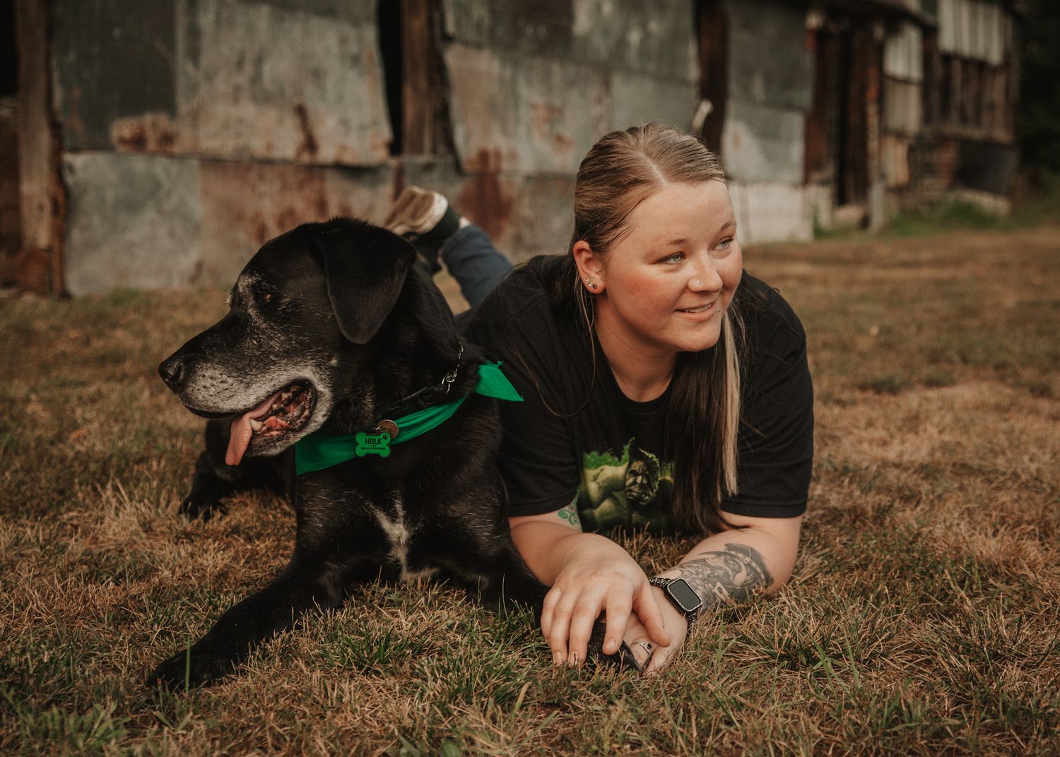 Woman with blonde hair lying on grass beside a black dog wearing a green bow tie outdoors.