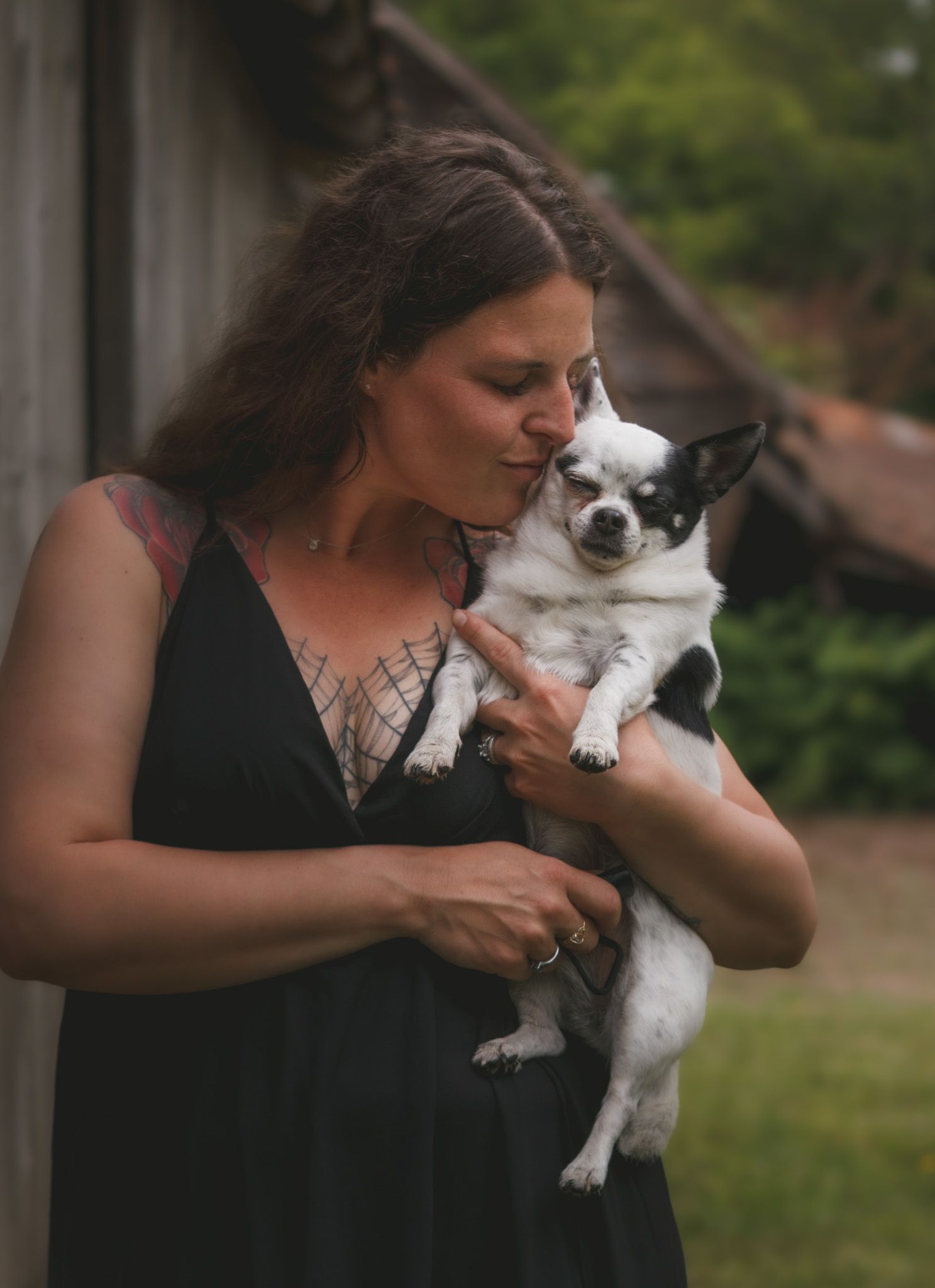 Woman in black dress kissing her black and white Chihuahua dog outdoors near a rustic building.