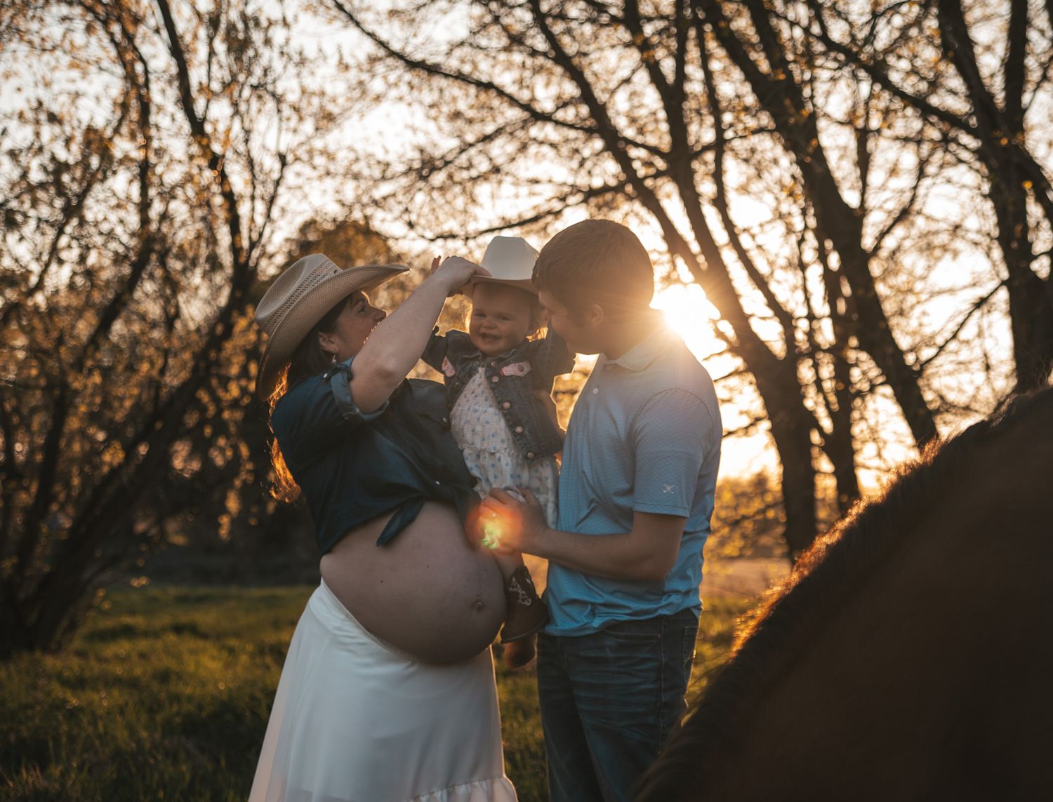 Pregnant couple sharing a tender moment at golden sunset, with the man lifting a toddler in a cowboy hat.