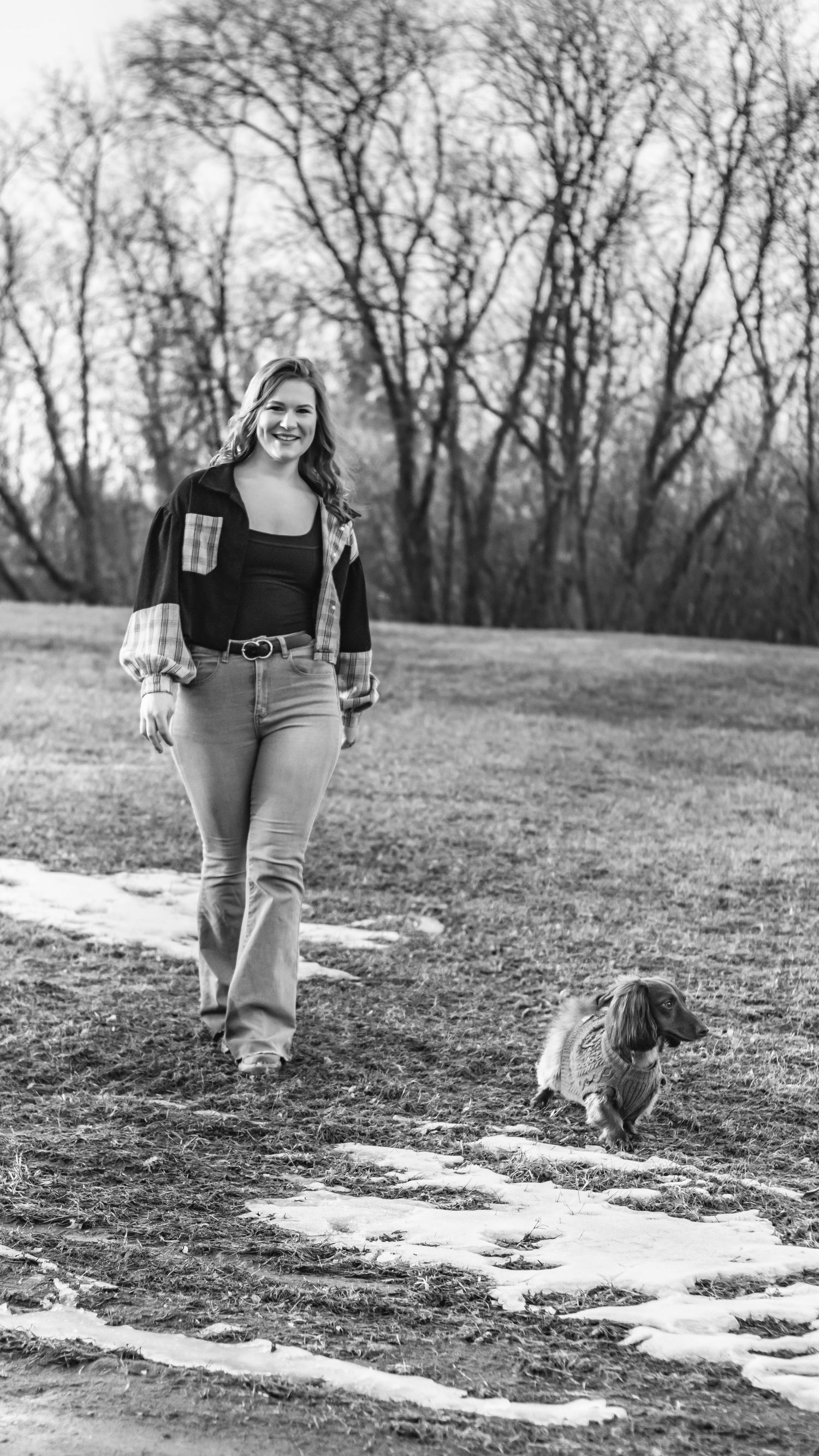 Woman in flannel shirt walking with a small dachshund dog in a black and white park photo.