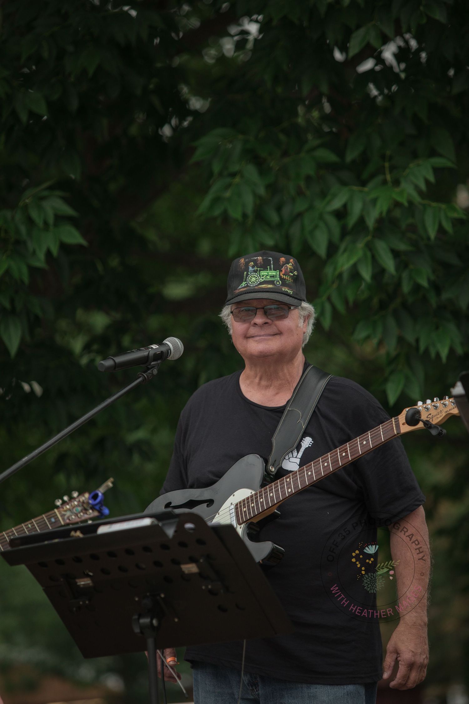 A guitarist in a black t-shirt and hat performs at an outdoor venue with stage equipment visible.