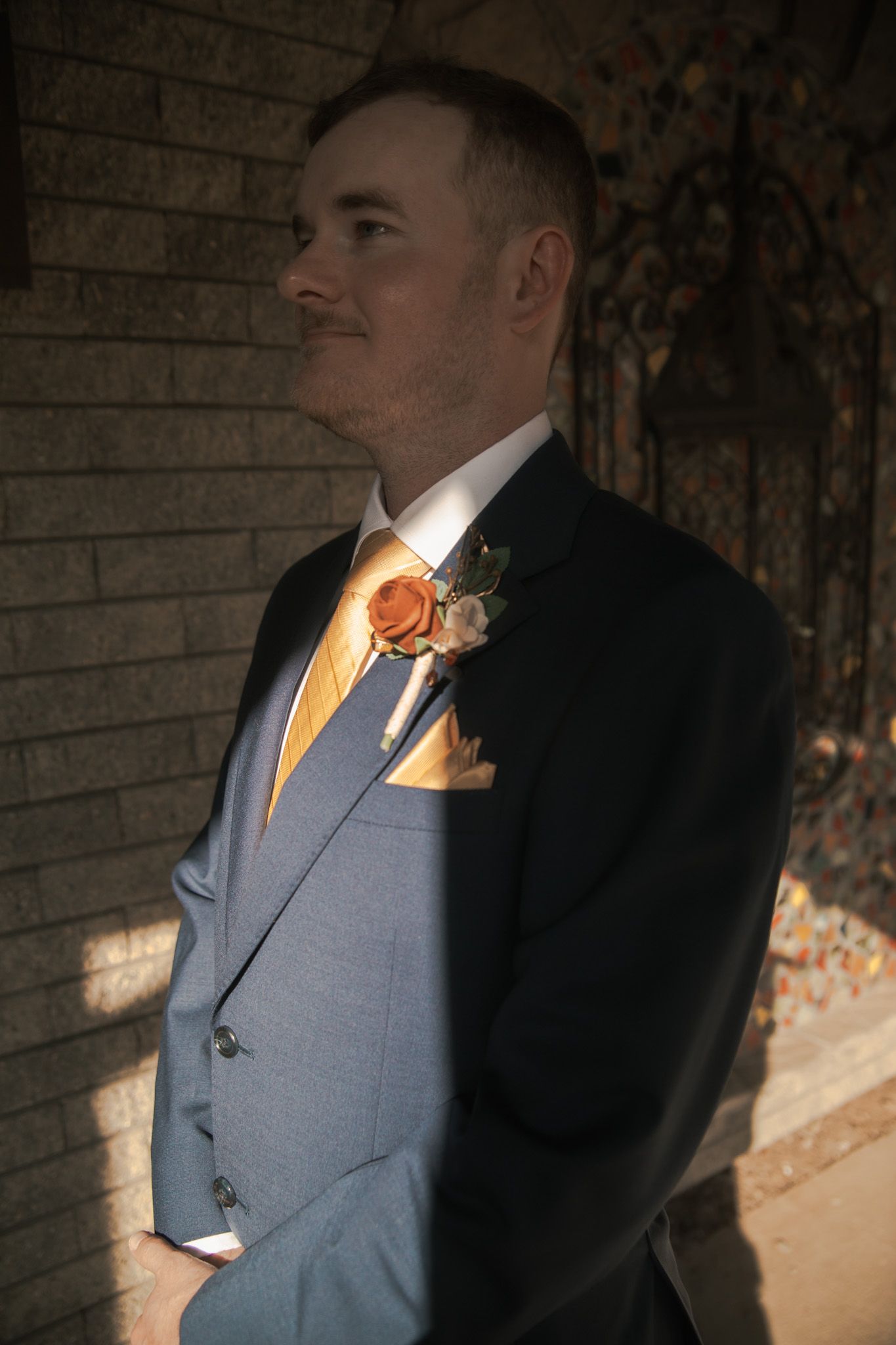Groom in grey suit with orange tie and boutonniere poses against rustic wooden wall in natural lighting.