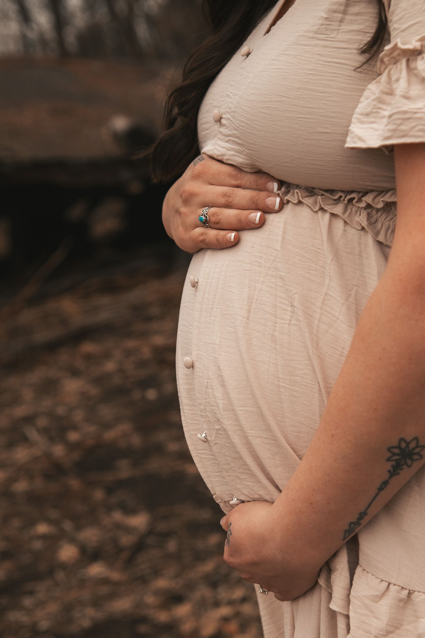 Close-up of pregnant woman in beige dress cradling baby bump outdoors, wearing turquoise ring.