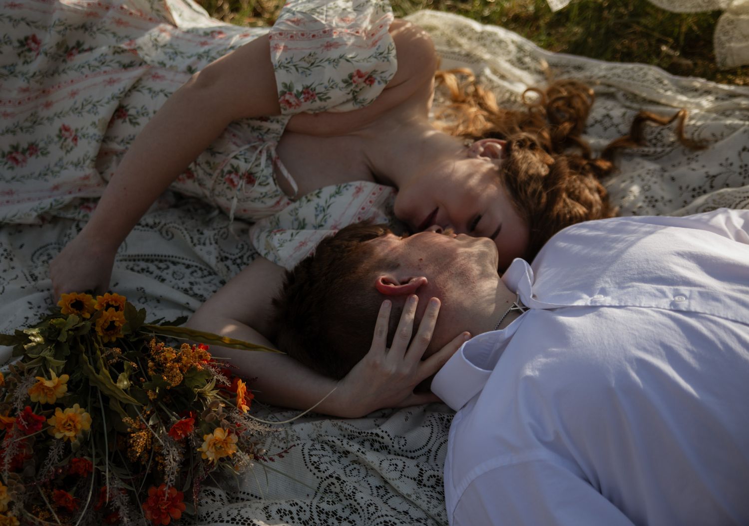 Romantic couple lying on a floral blanket outdoors, sharing an intimate moment with wildflowers nearby.