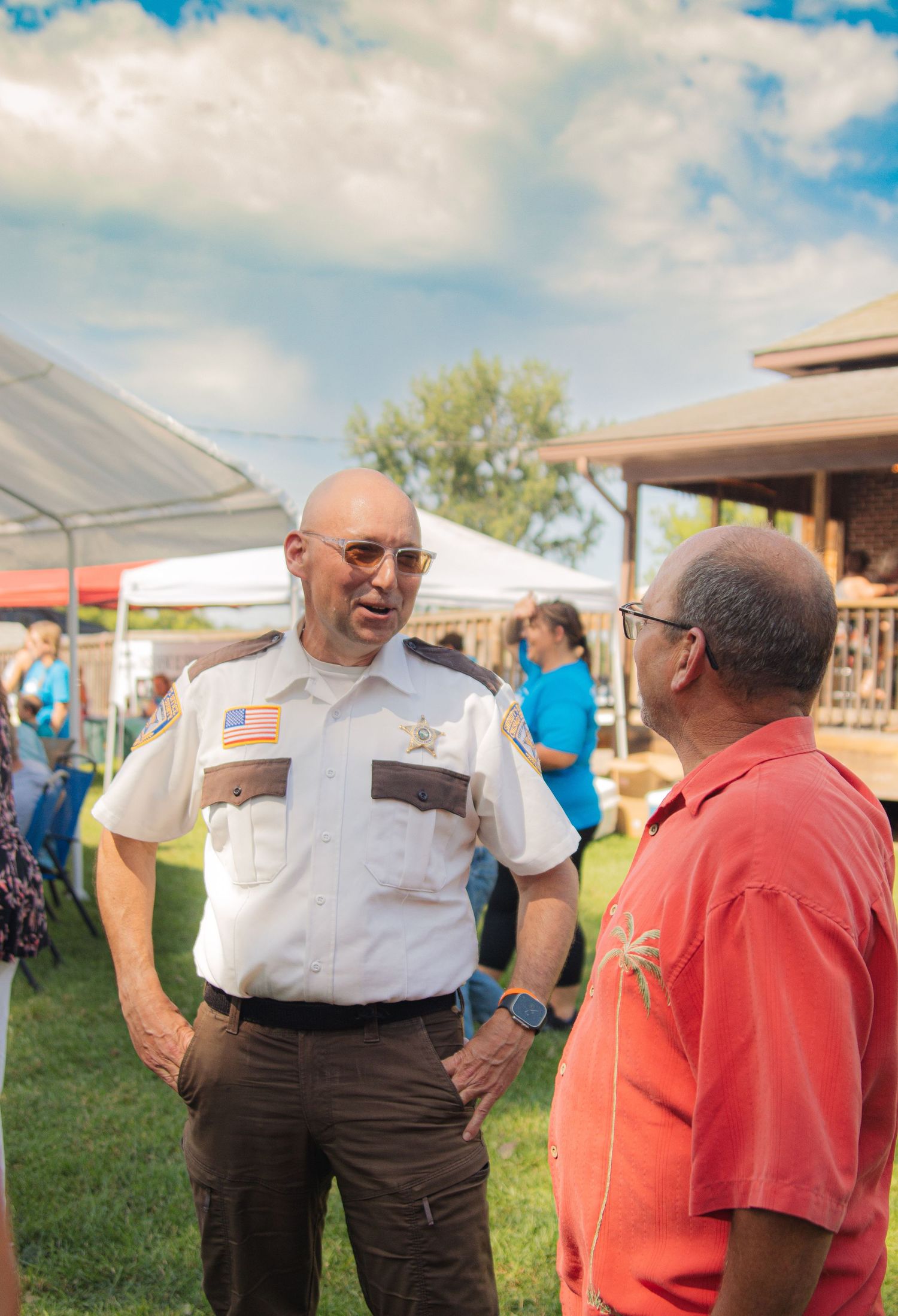 People having an animated conversation at an outdoor community event on a sunny day with white tents visible.