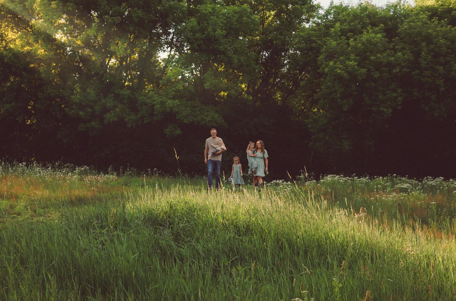 Family of three standing in a lush green meadow surrounded by tall trees in warm natural light.