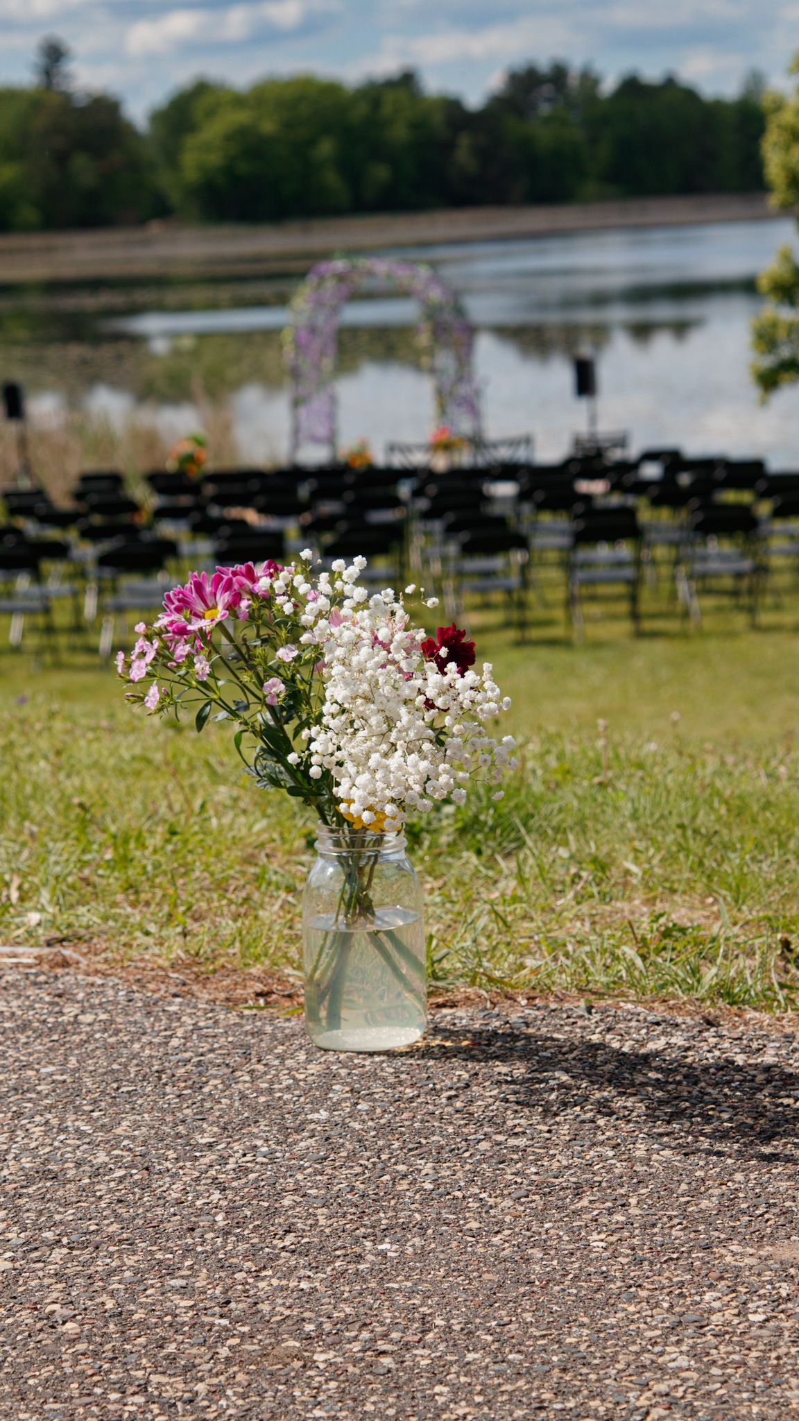 Multiple mason jars with white and pink wildflowers line a gravel path near a tranquil lakeside setting.