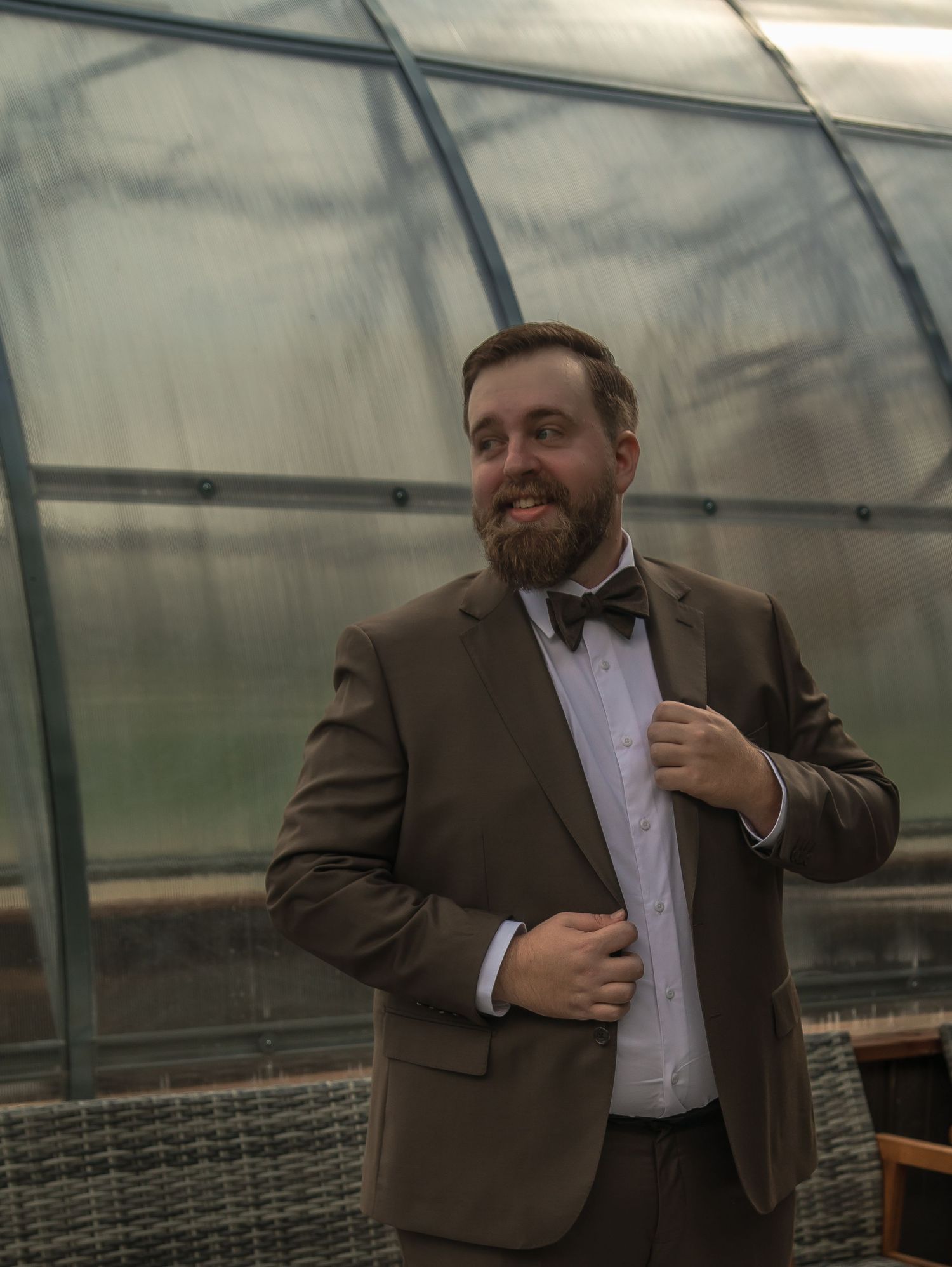 A person in a brown suit jacket adjusts their collar while standing in a greenhouse setting.