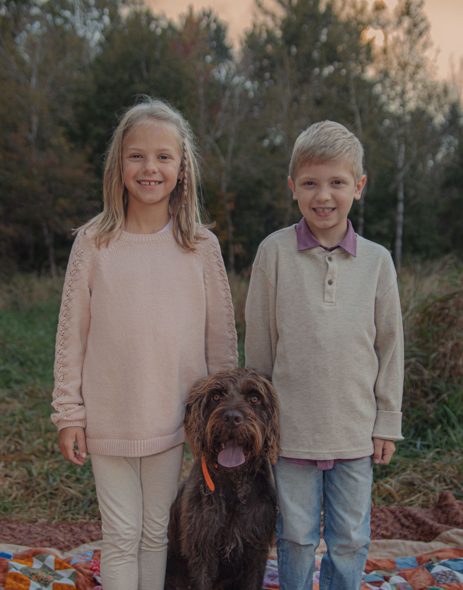 Two children and a large brown shaggy dog pose together on a colorful blanket during an autumn outdoor photoshoot.