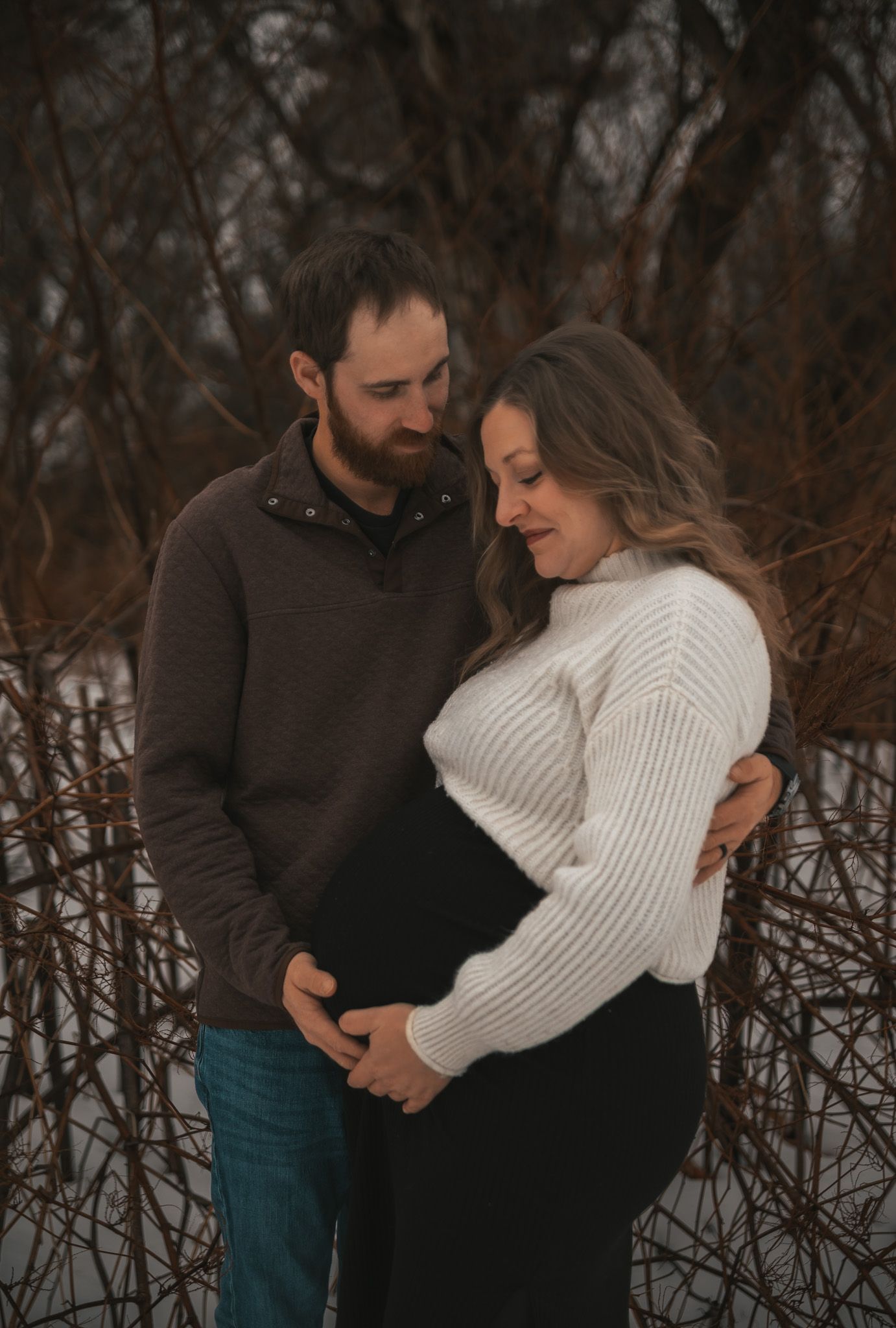 A romantic winter photoshoot showing a couple embracing in a snowy forest setting with bare branches in the background.