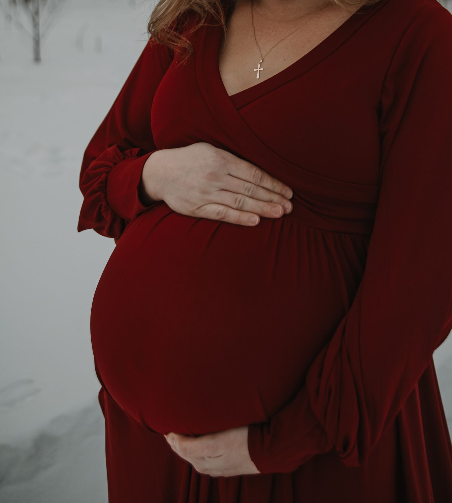 Woman in burgundy dress cradles her pregnant belly against snowy winter backdrop.