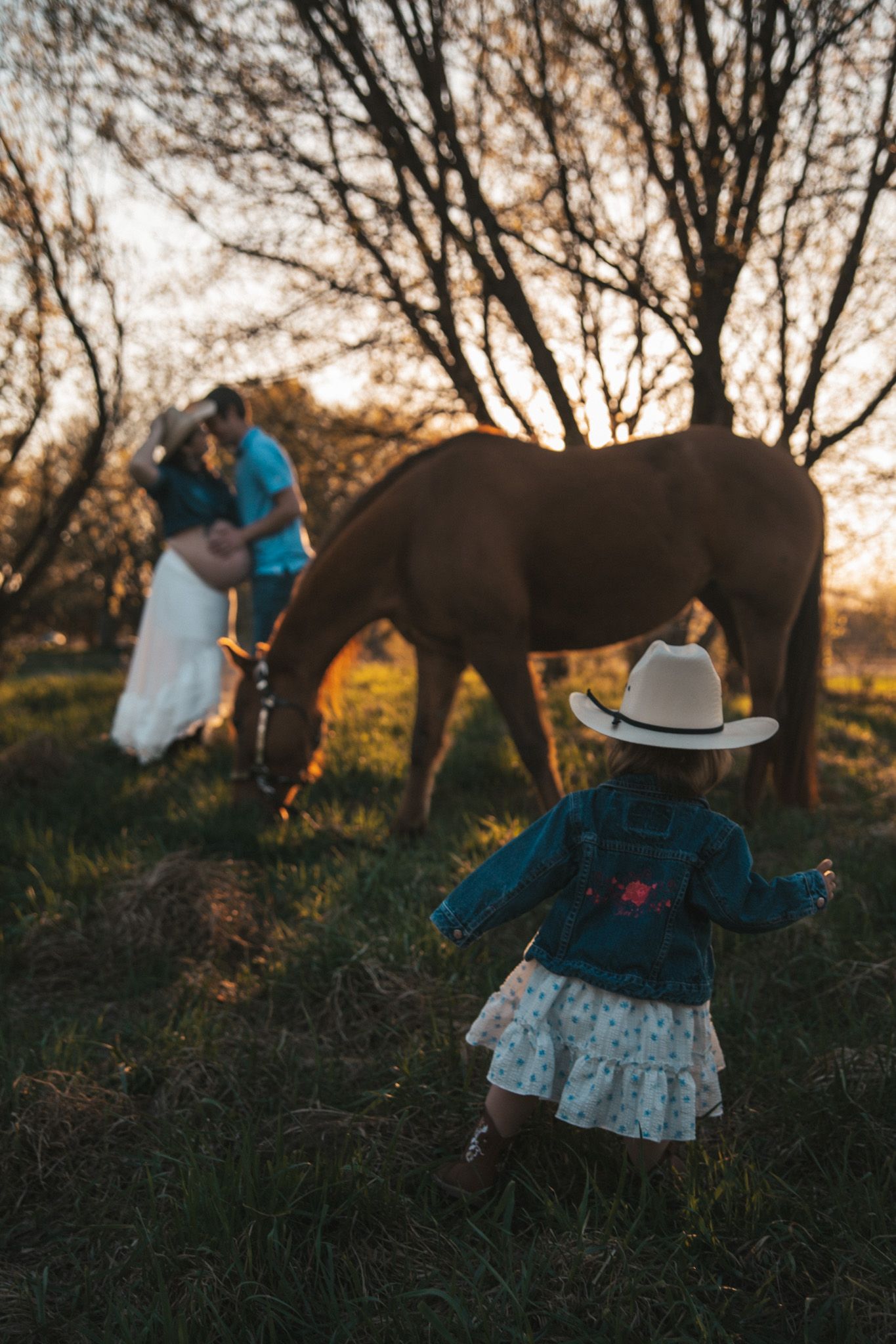 Little girl in cowboy hat and denim jacket walks toward parents kissing near grazing horse at sunset.