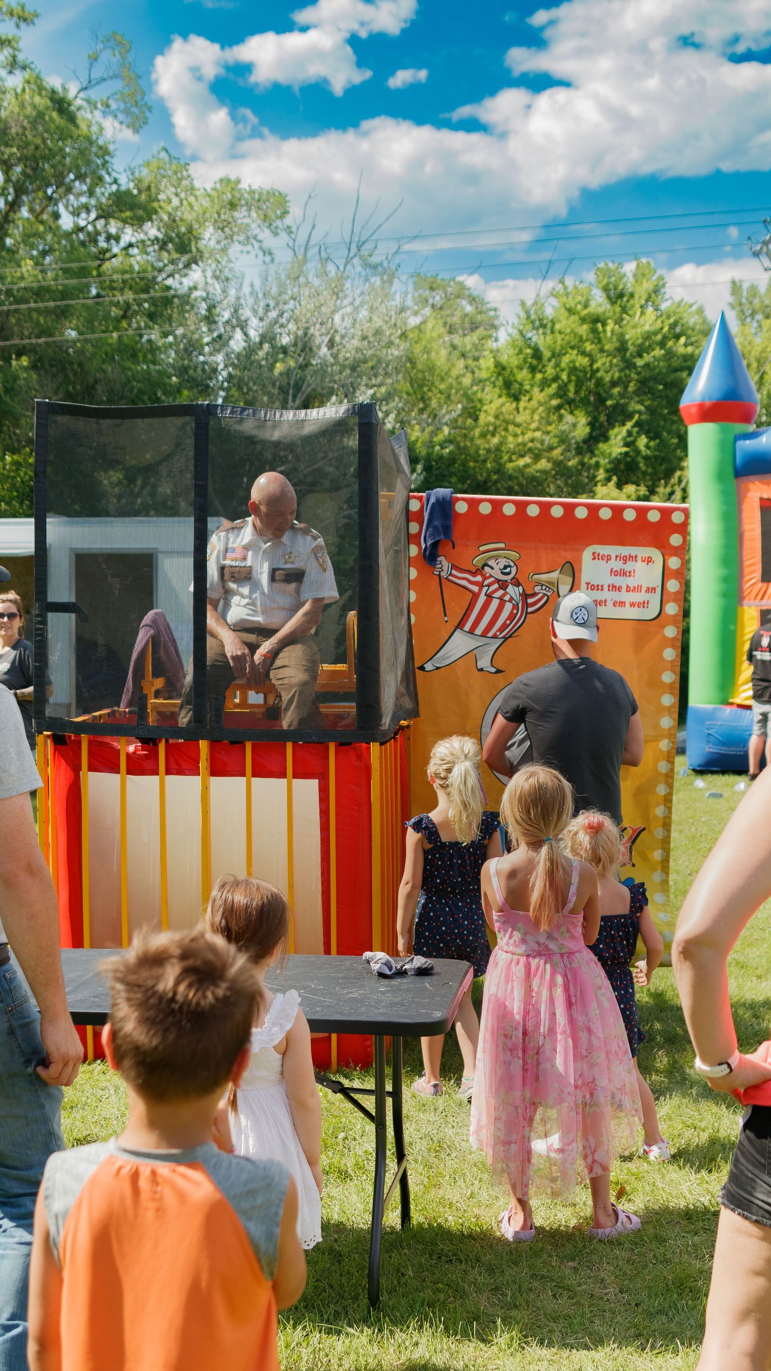 Children watch a dunk tank attraction at an outdoor carnival with bounce house and decorations.