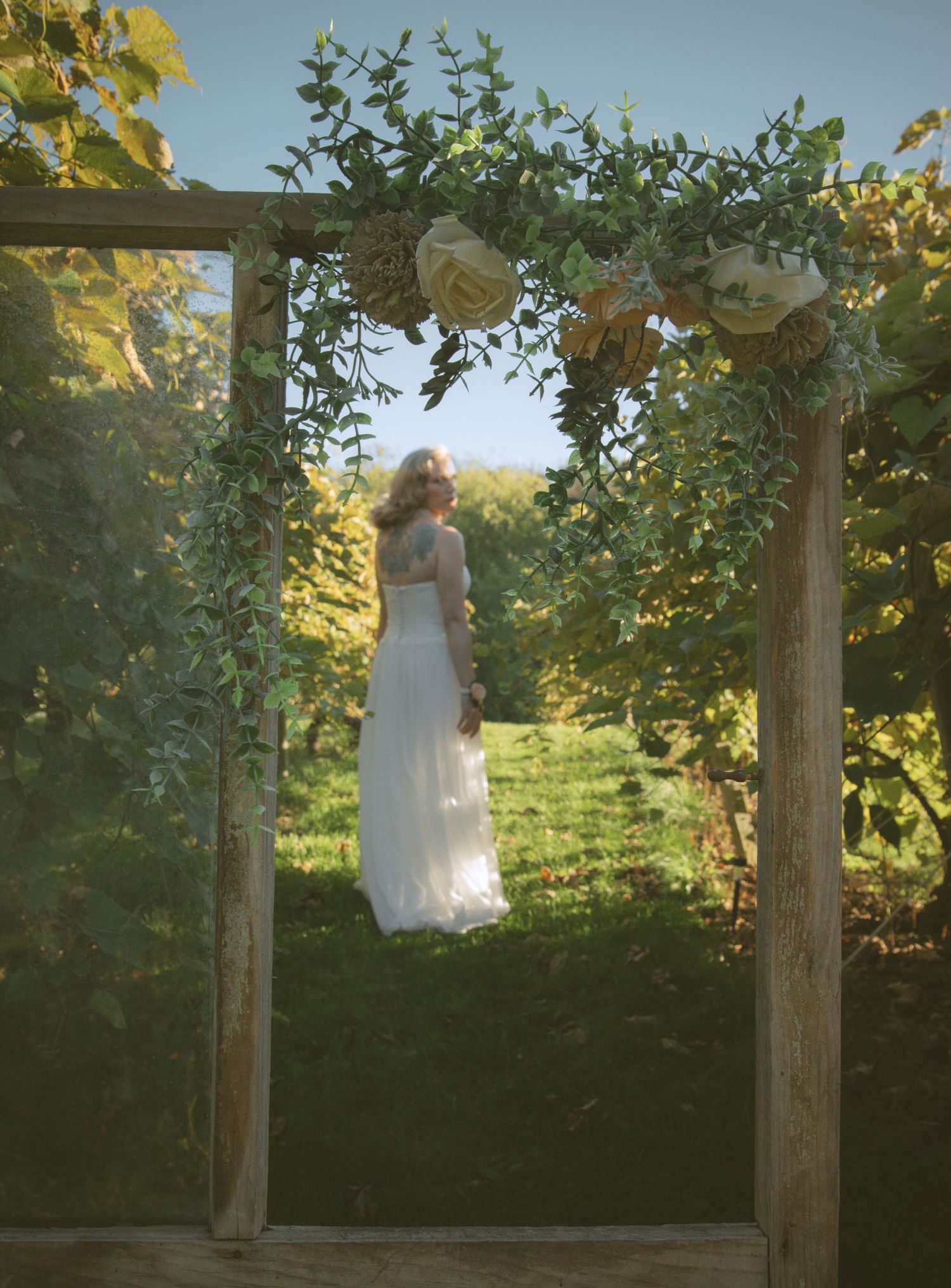 A dreamy wedding arch decorated with white roses frames a sunlit garden path during golden hour.