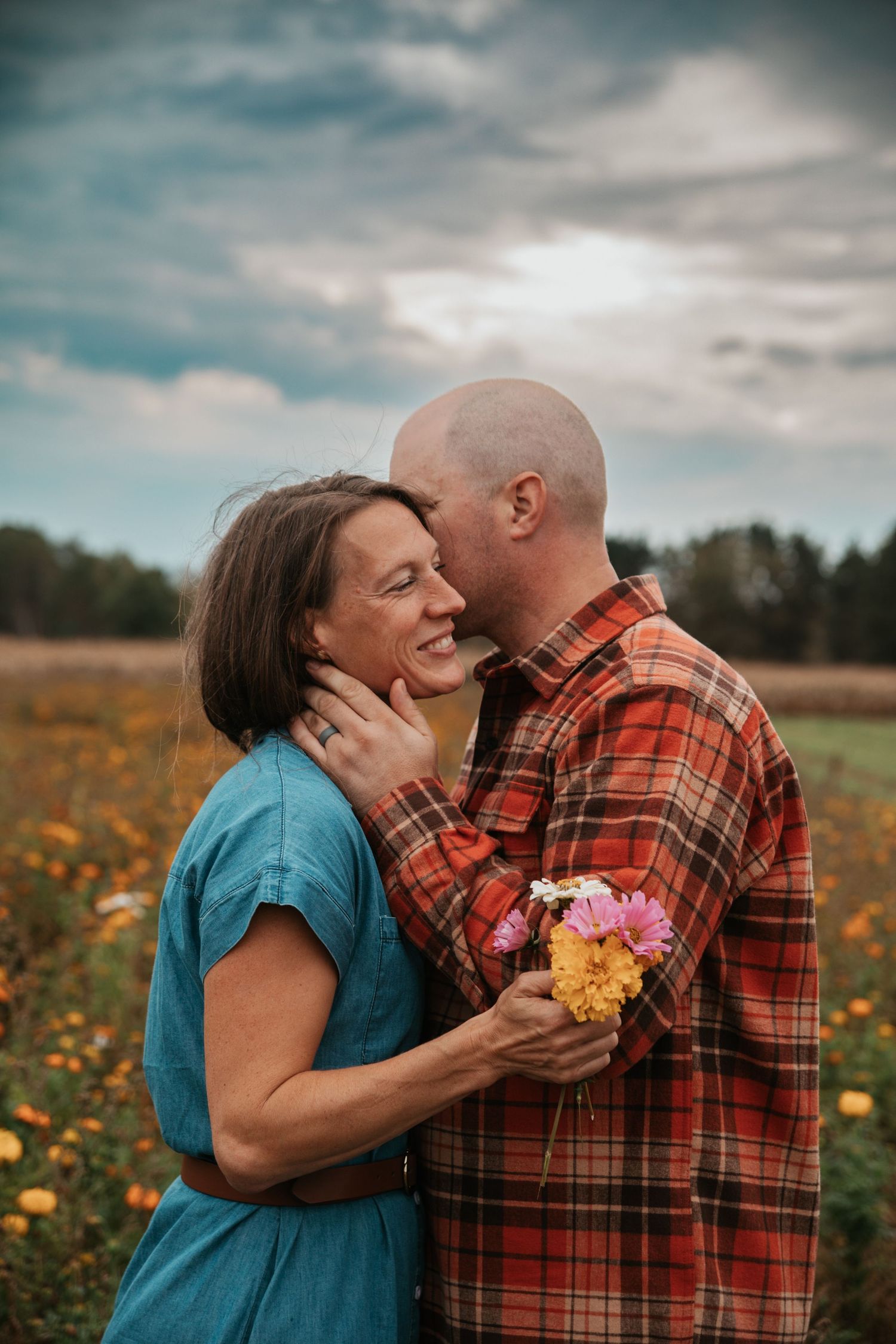 A couple shares a tender moment in a wildflower meadow, wearing a plaid shirt and teal top against a cloudy sky.