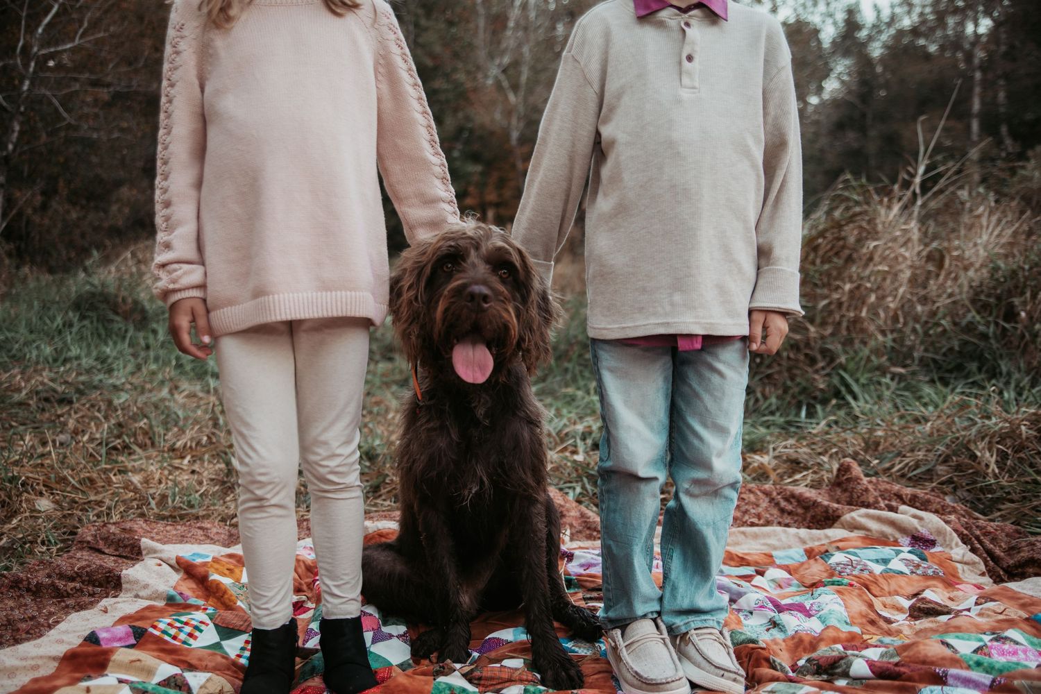 Two people in neutral-colored outfits stand on a colorful patchwork quilt blanket outdoors in nature.