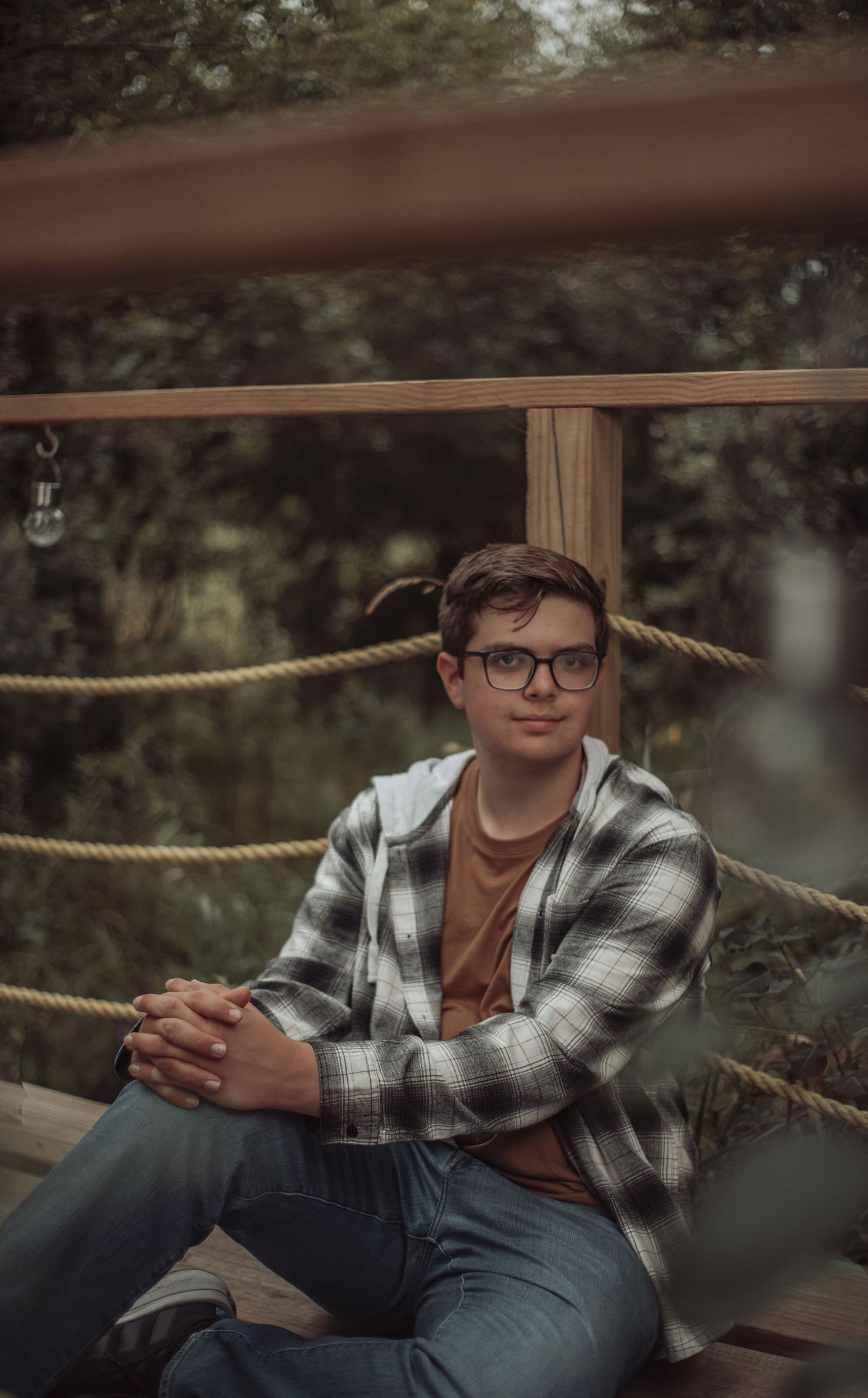 A person in glasses and plaid flannel shirt poses casually on wooden steps outdoors in a natural setting.