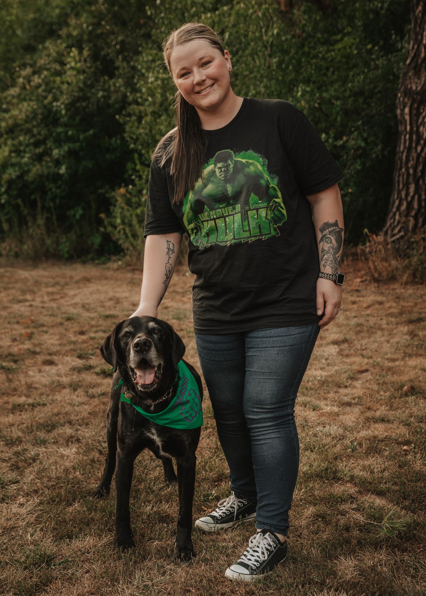 Smiling woman in a Hulk shirt standing with her black dog wearing a green bandana outdoors.