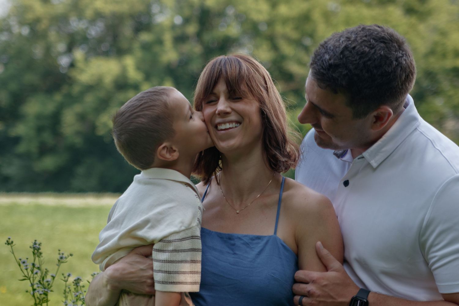 Family enjoys tender moments together outdoors on a sunny day in casual summer attire.
