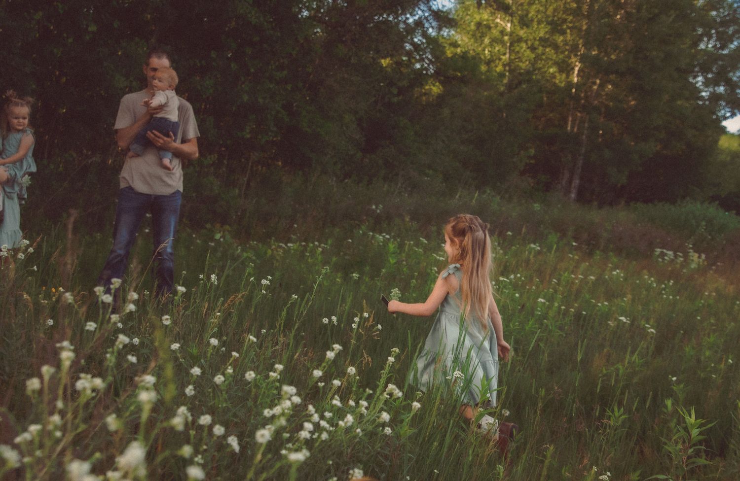 A young girl in a teal dress walks through a wildflower meadow as a man watches in a sunlit forest clearing.