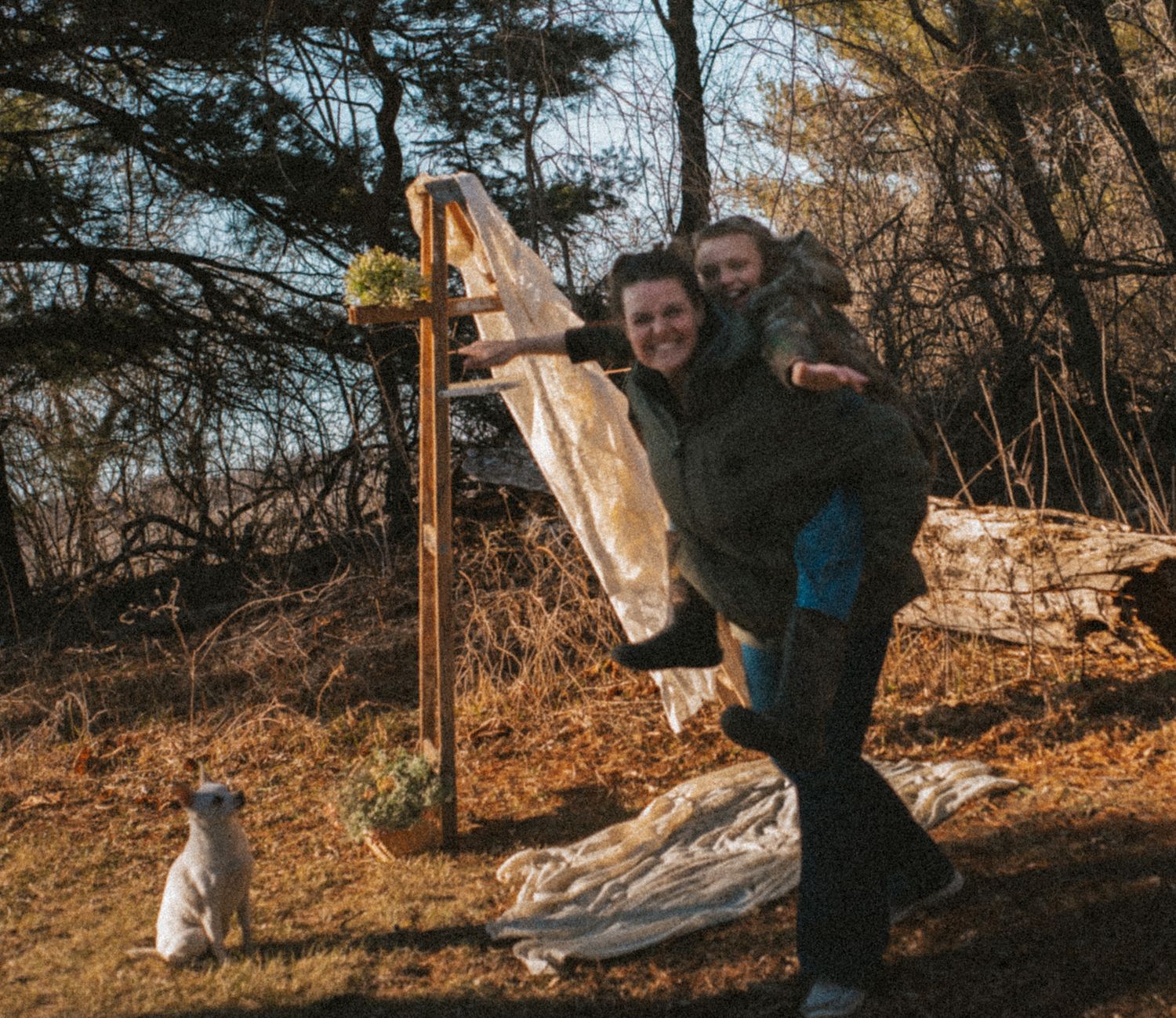 Two people laughing outdoors near a wooden cross and driftwood, surrounded by trees in a natural setting.