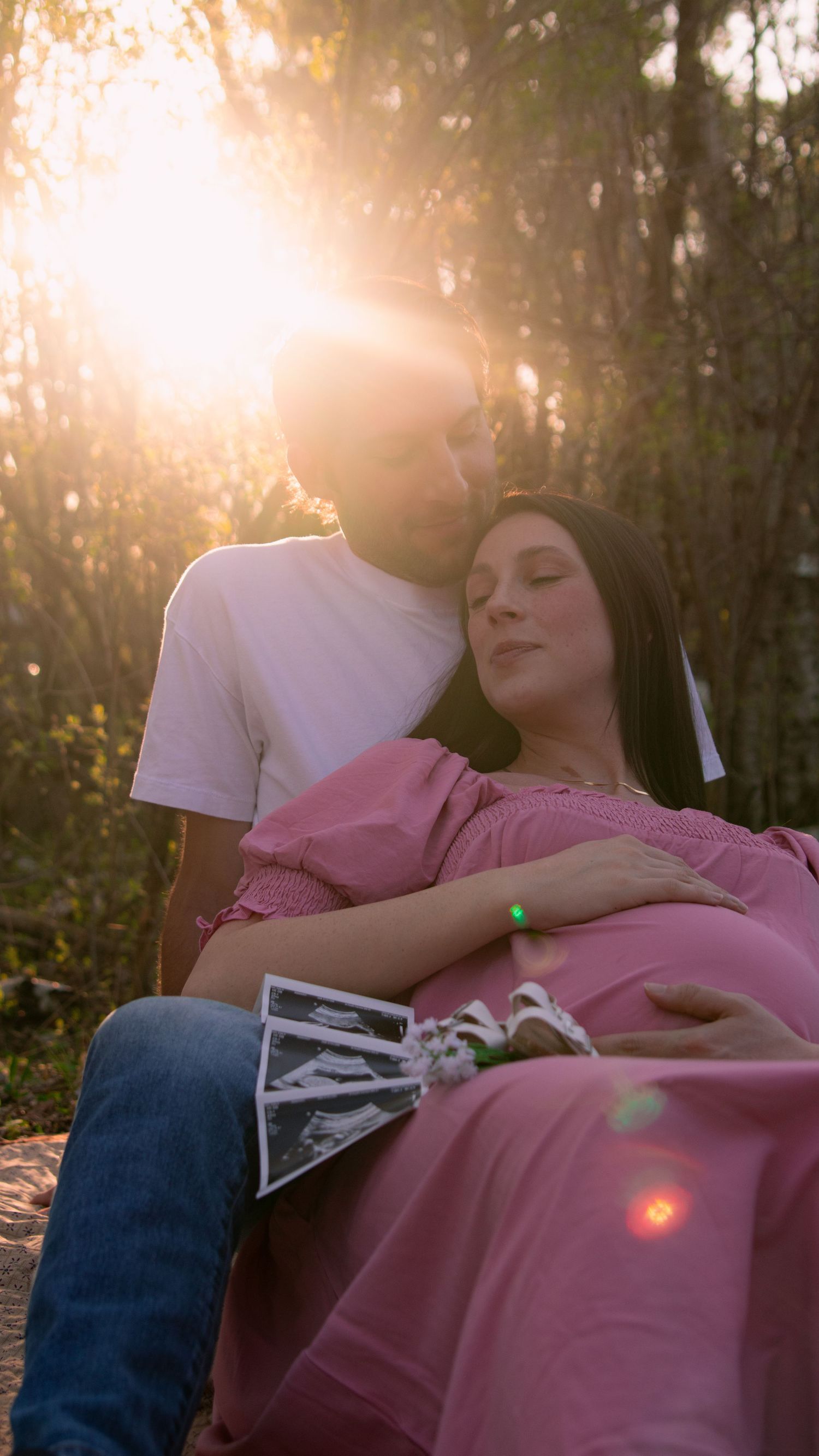 A couple rests together outdoors at sunset while holding a sonogram photo in a tender moment.