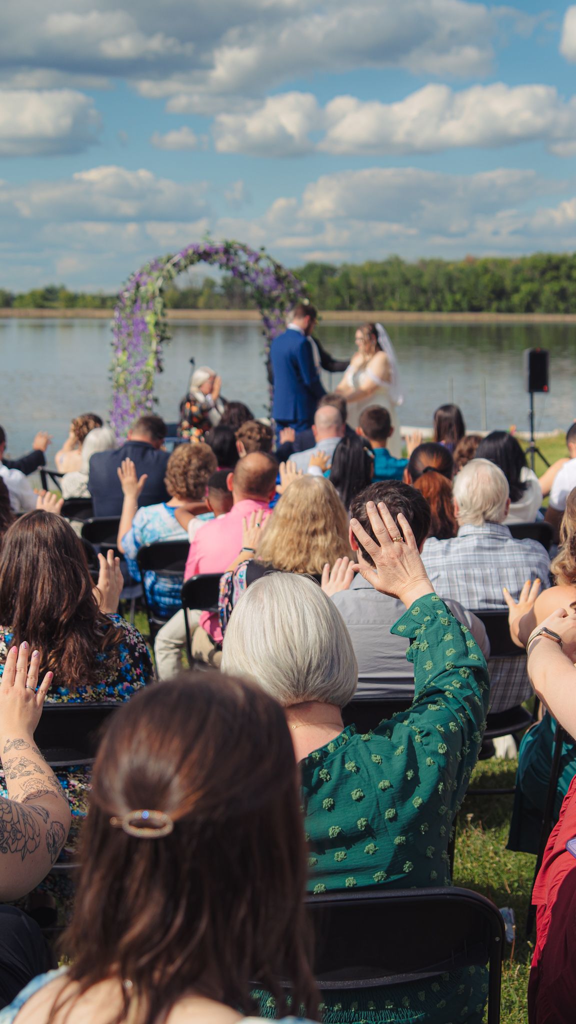 Outdoor wedding ceremony by a lake with guests seated in chairs on a grassy lawn under blue skies.