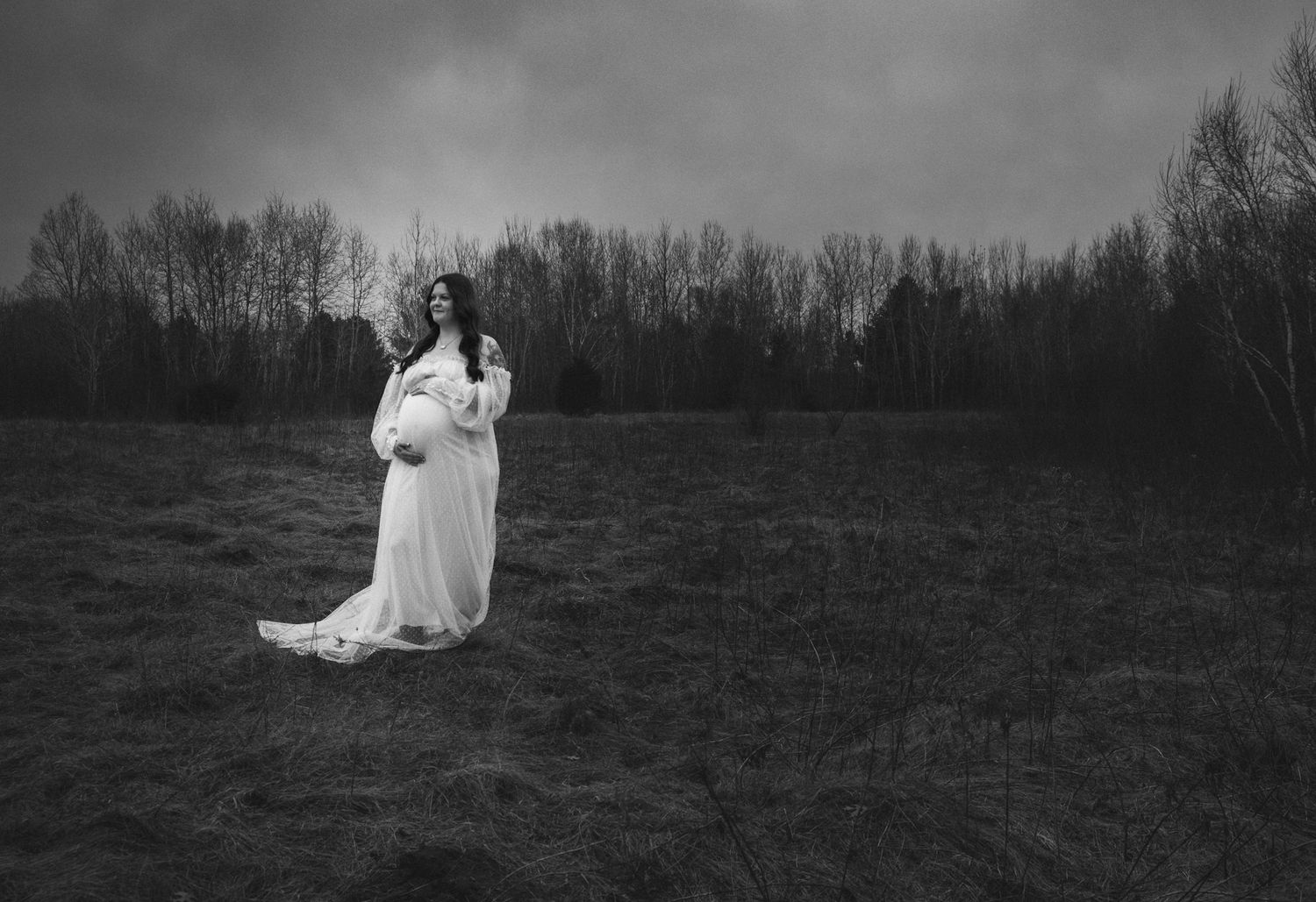 Black and white maternity photo of pregnant woman in flowing white gown standing in open field.
