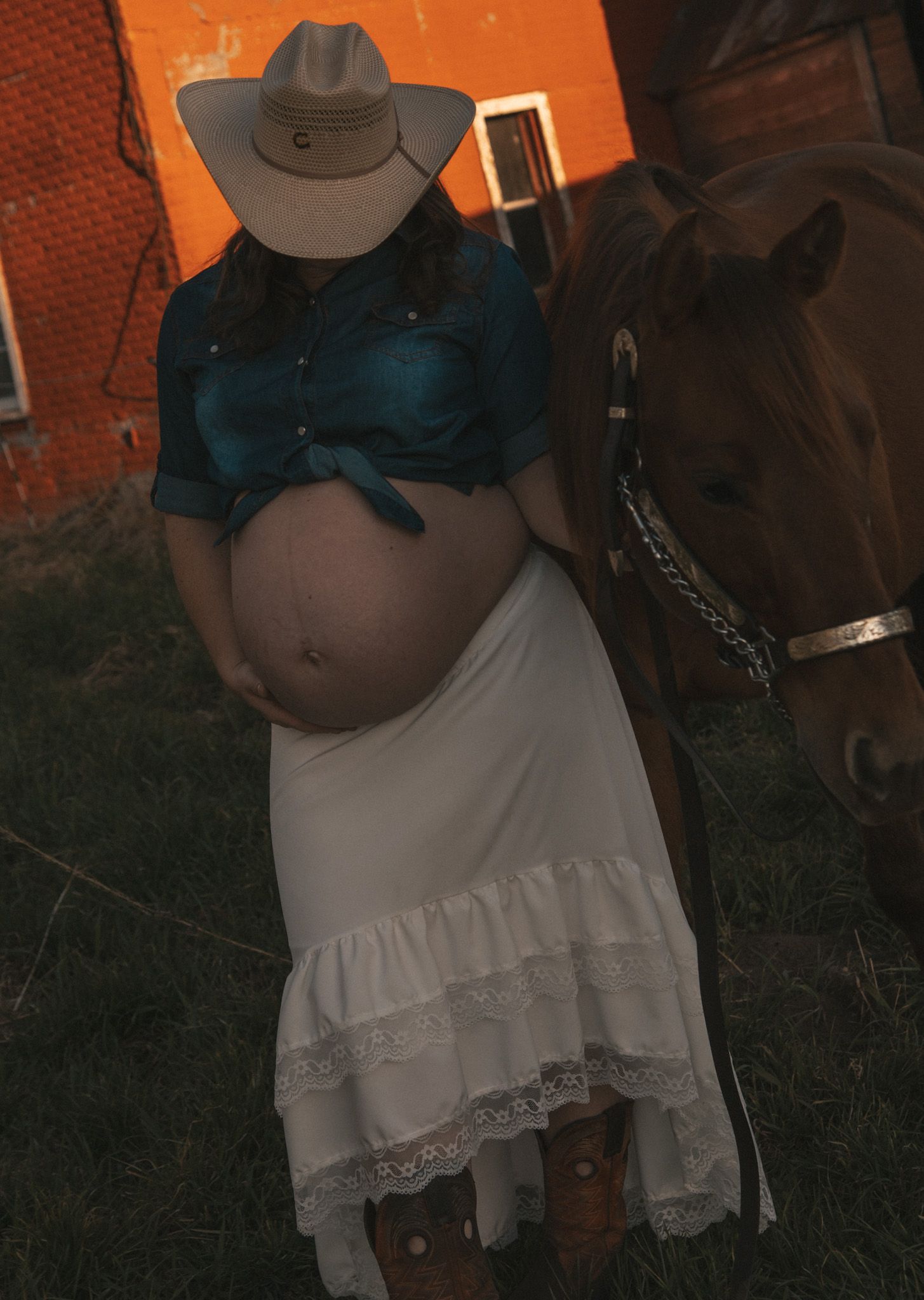 Pregnant woman in cowboy hat, blue denim shirt, white ruffled skirt, and boots poses beside a chestnut horse near a brick wall.