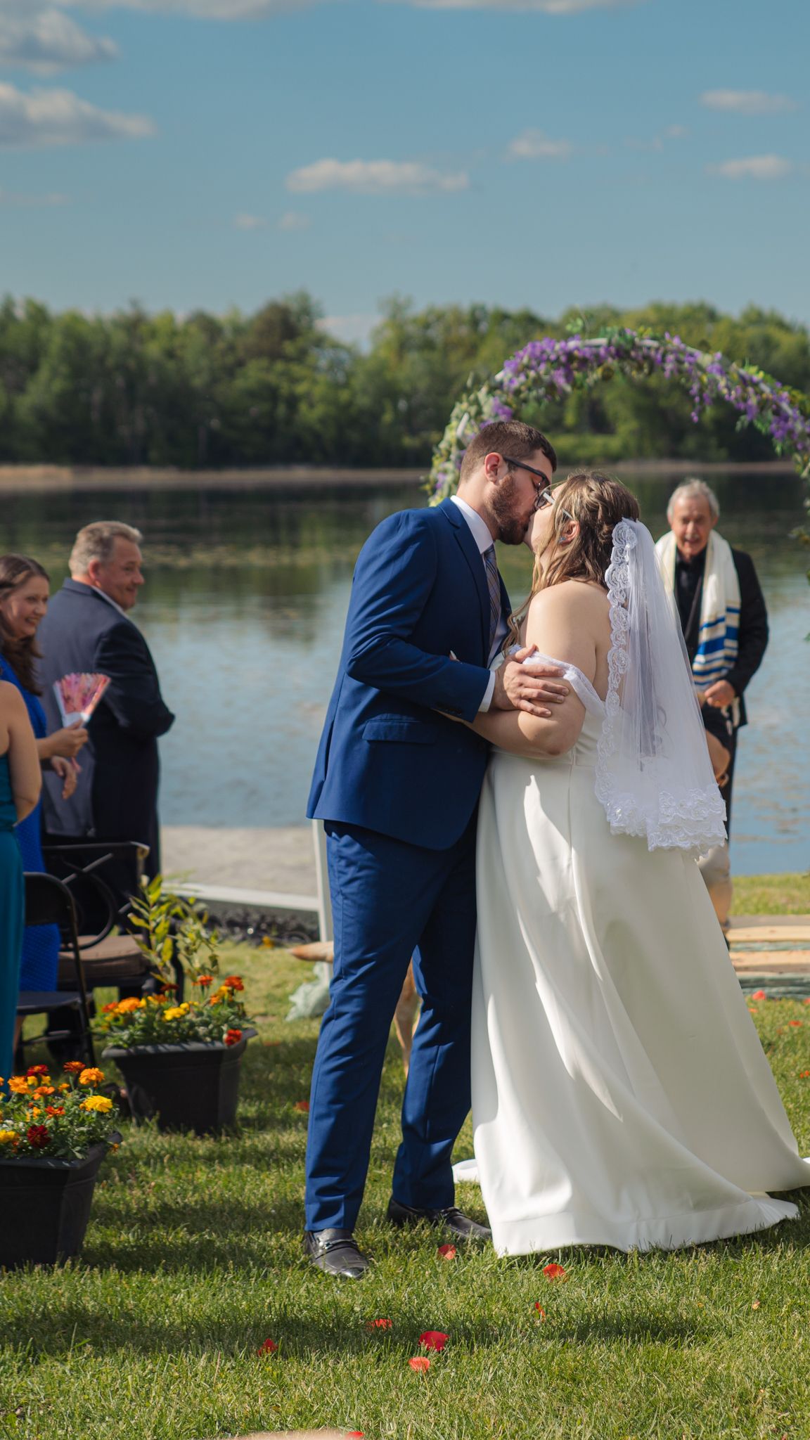 A lakeside wedding ceremony takes place on a sunny day with a floral arch and orange flowers decorating the green lawn.
