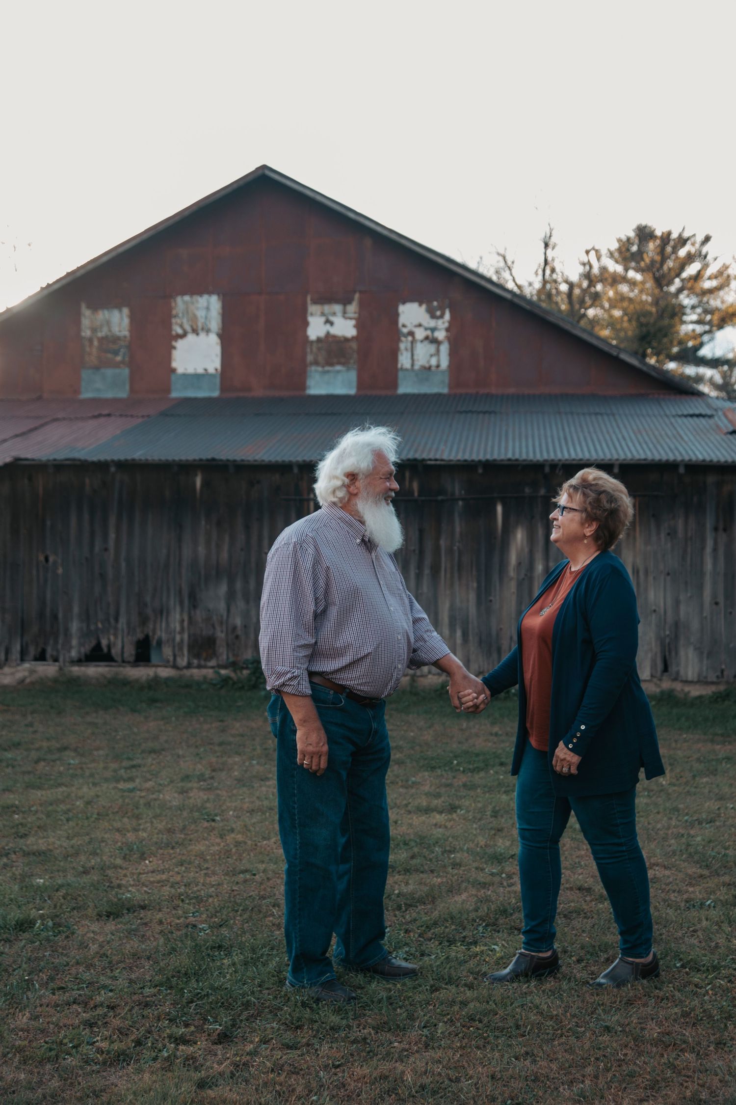 An elderly couple stands together in front of a rustic red barn on a grassy field at dusk.