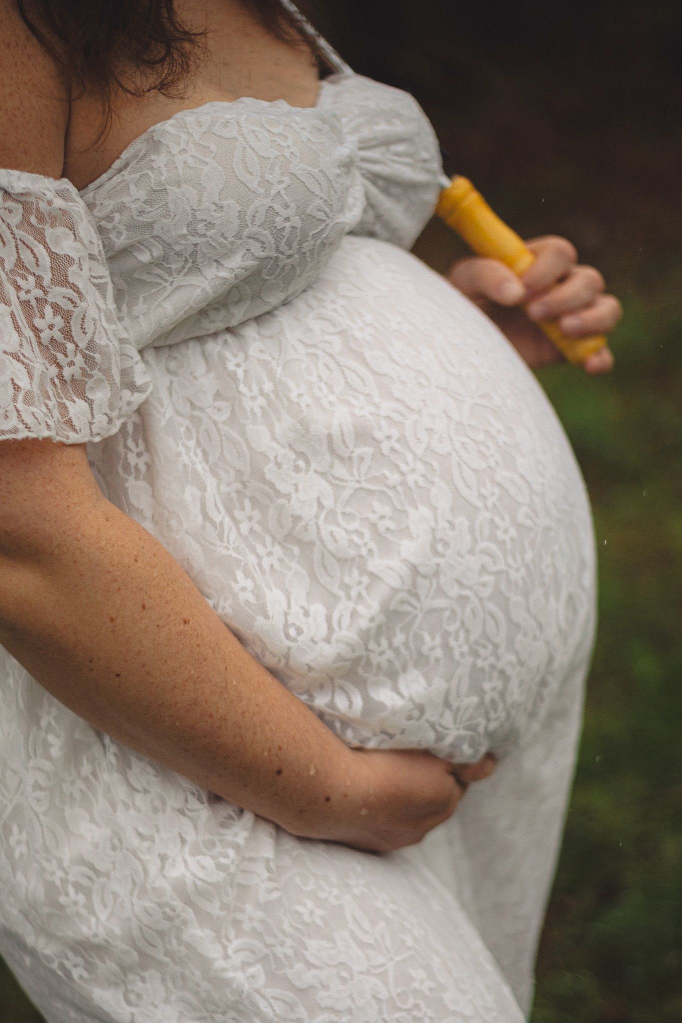 Pregnant woman in white lace off-shoulder dress cradling baby bump outdoors.